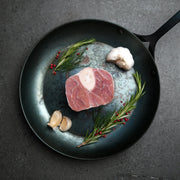 Milk Fed Dutch Veal Osso Buco Shank in skillet with garlic, rosemary, and red peppercorns, showcasing its raw, bone-in cut ready for braising.