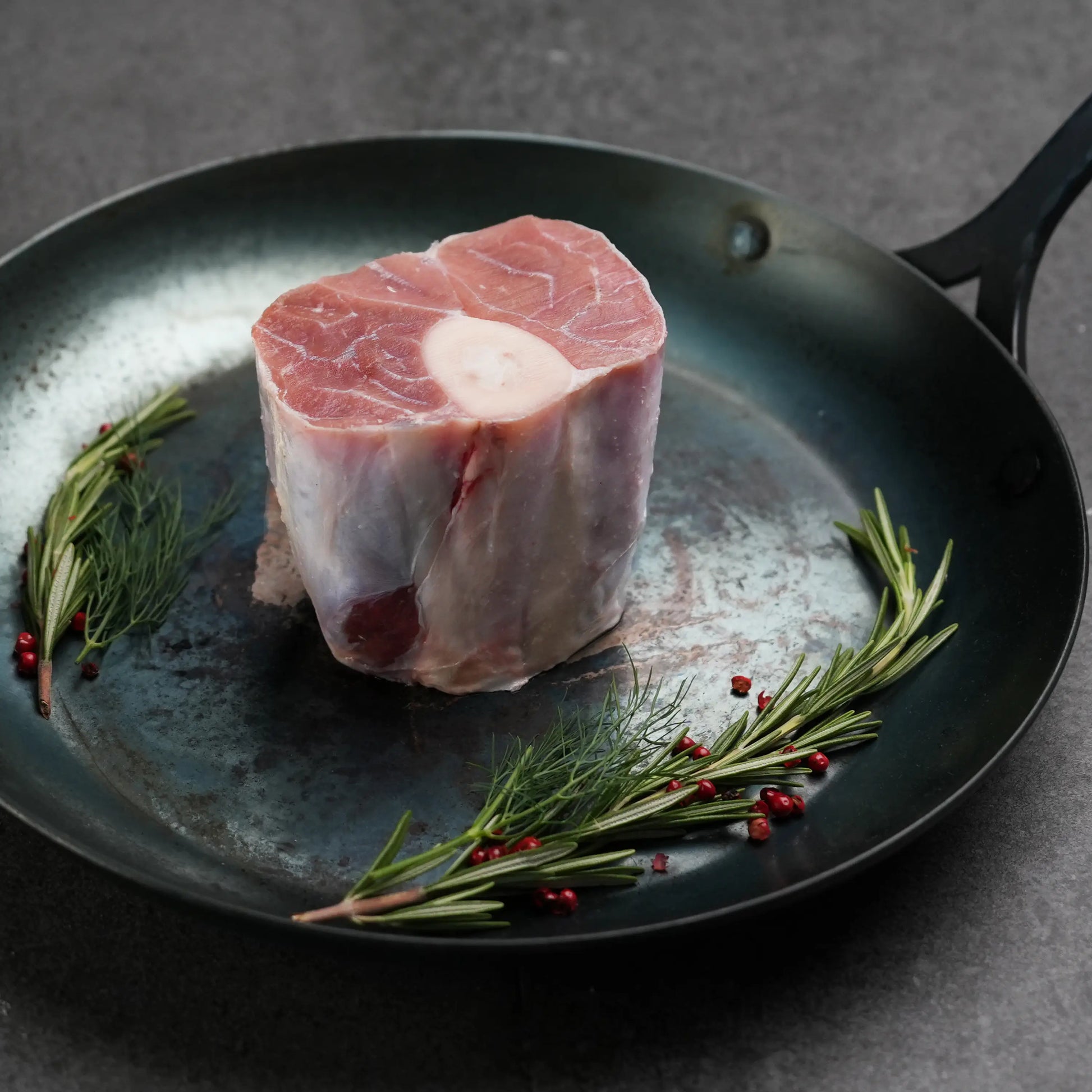 Milk Fed Dutch Veal Osso Buco Shank in metal pan with rosemary and red peppercorn garnish, raw and ready for slow cooking and braising.