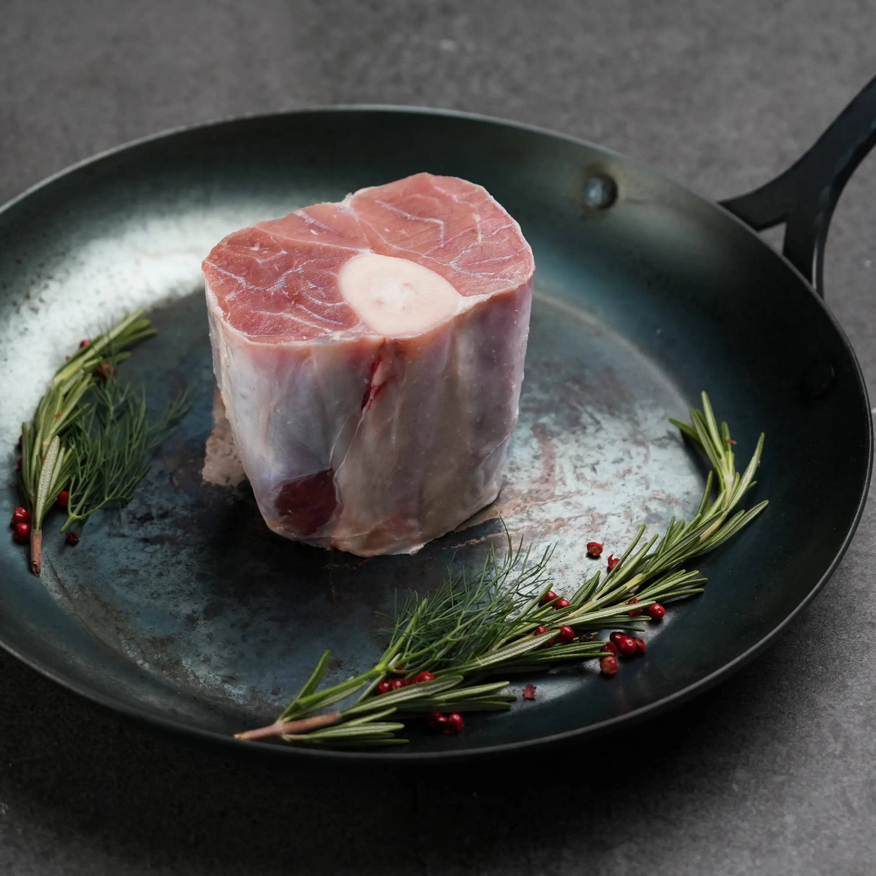 Milk Fed Dutch Veal Osso Buco Shank in metal pan with rosemary and red peppercorn garnish, raw and ready for slow cooking and braising.