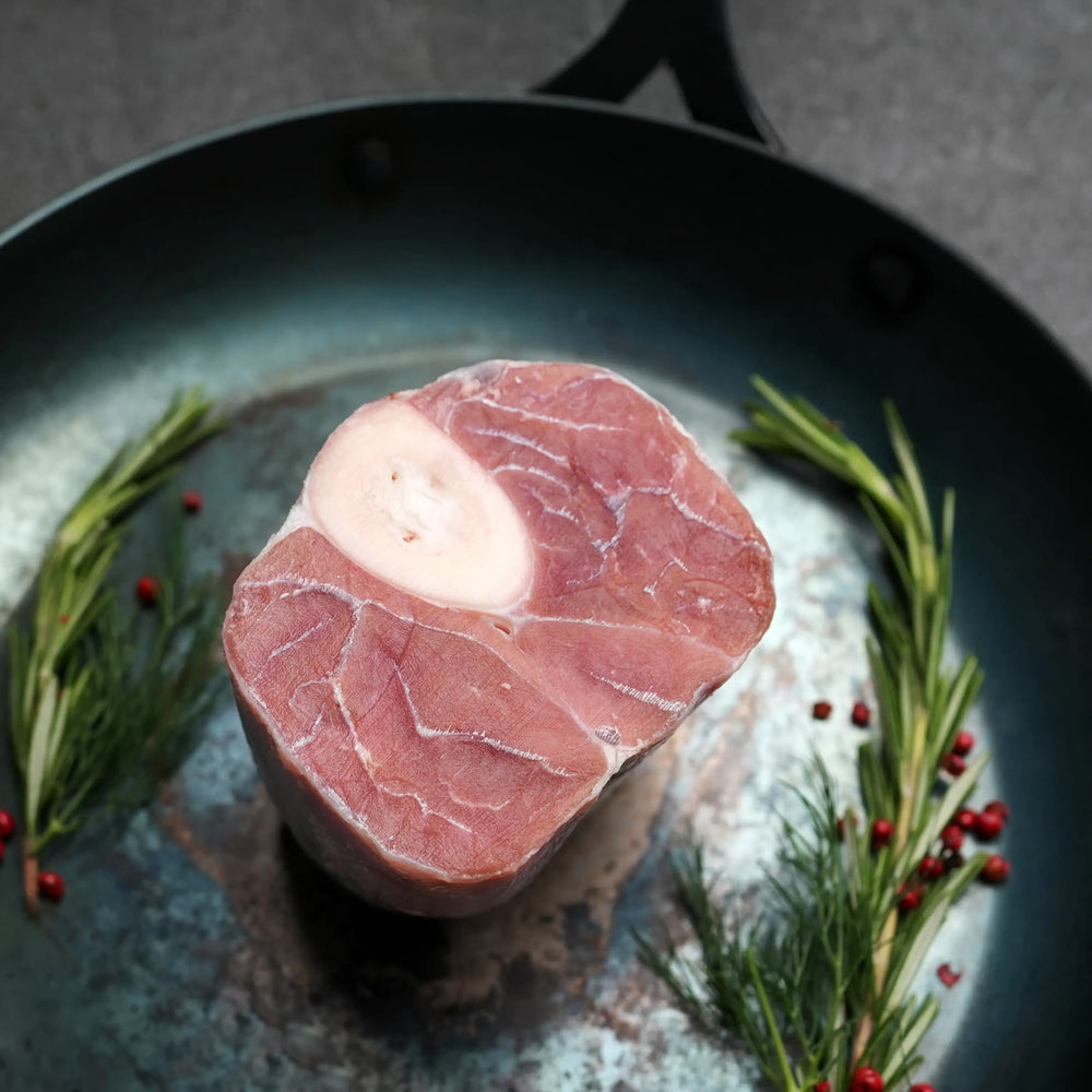 Milk Fed Dutch Veal Osso Buco Shank in a metal pan with fresh rosemary sprigs and red peppercorns, ready for slow cooking to tender perfection.