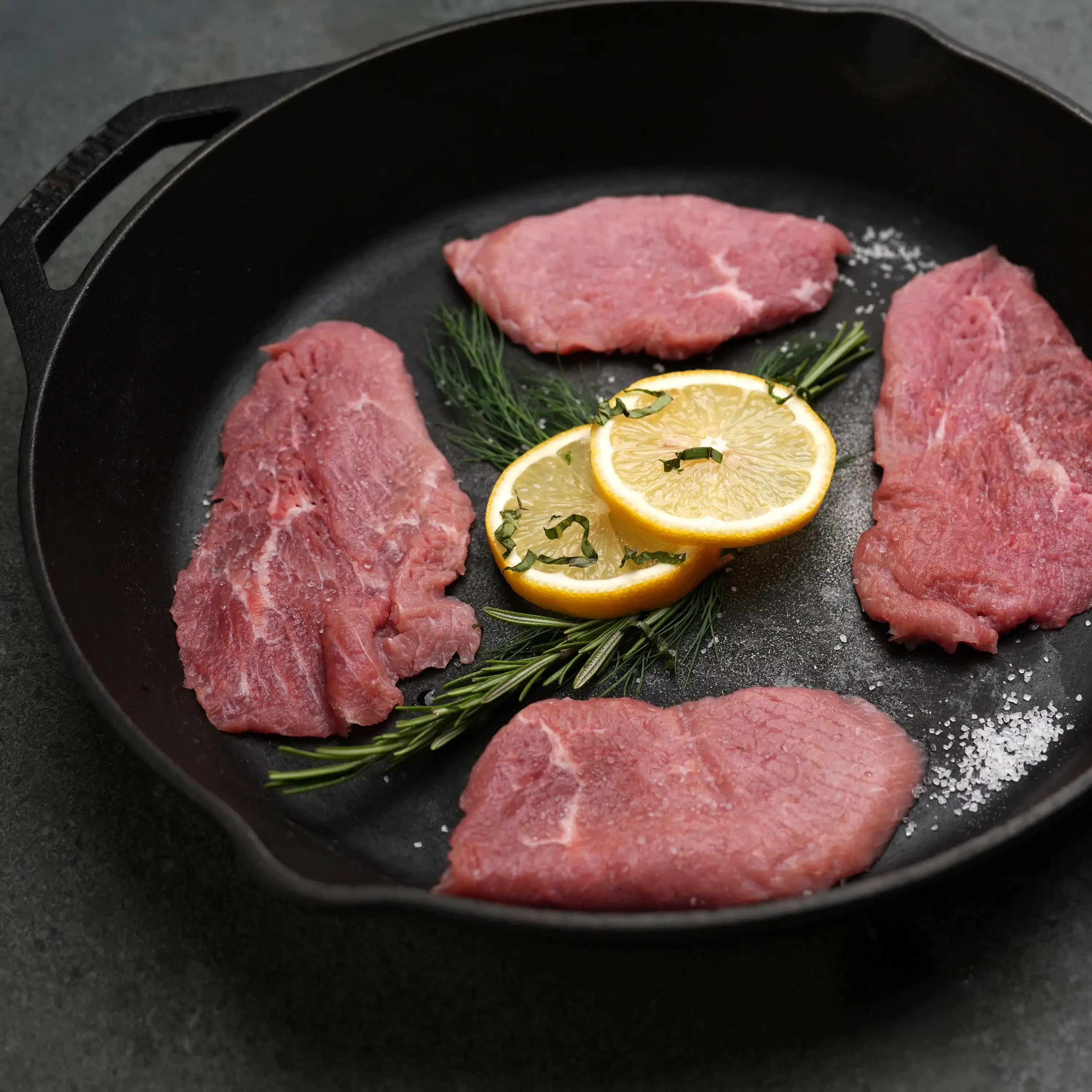 Milk Fed Dutch Veal Scallopine Slices in a cast-iron skillet with fresh herbs and lemon slices, showcasing tender, hormone-free veal ready for cooking.