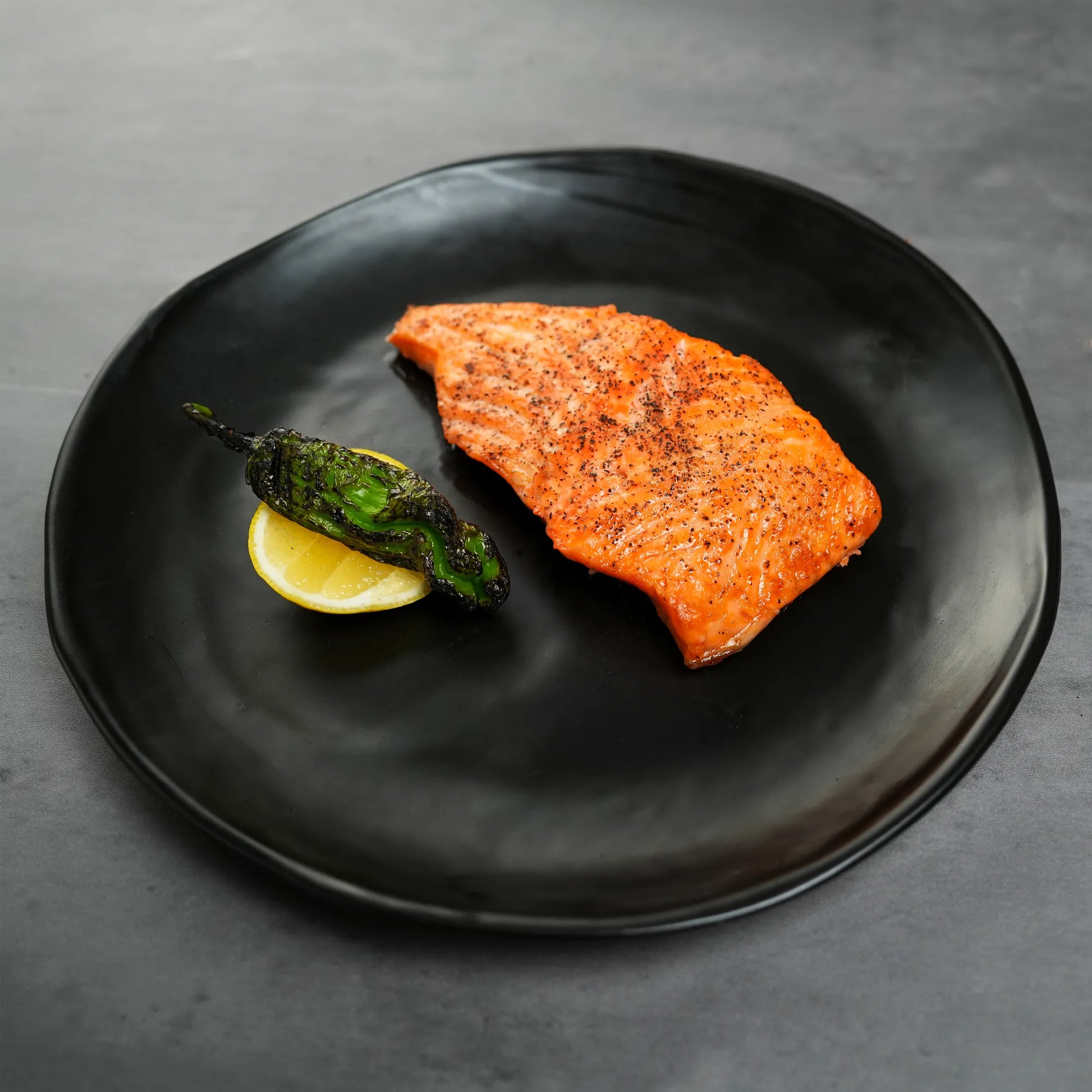 Salmon fillet with a lemon wedge and green herb on a black plate against a gray background