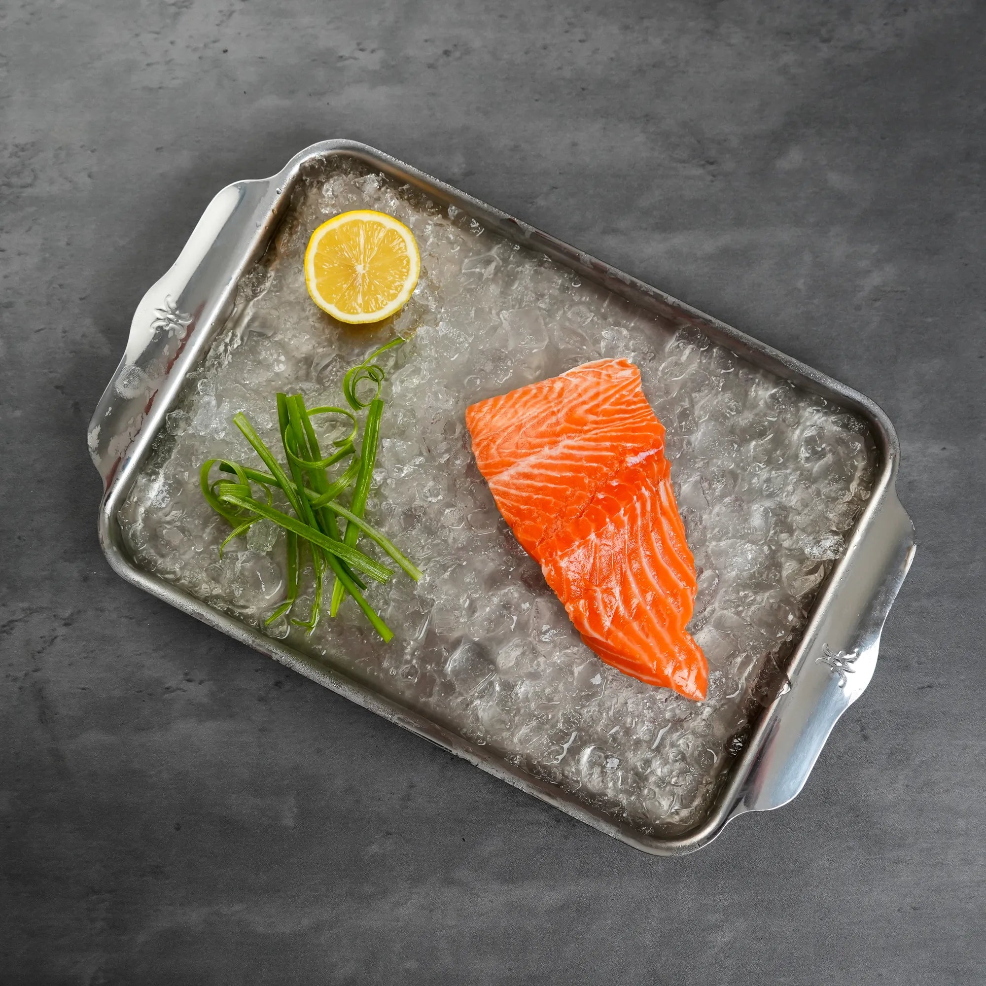 Raw salmon fillet on a bed of ice with a lemon and green onions on a dark surface