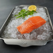 Salmon fillet on ice with a lemon wedge and greenery in a metal tray.