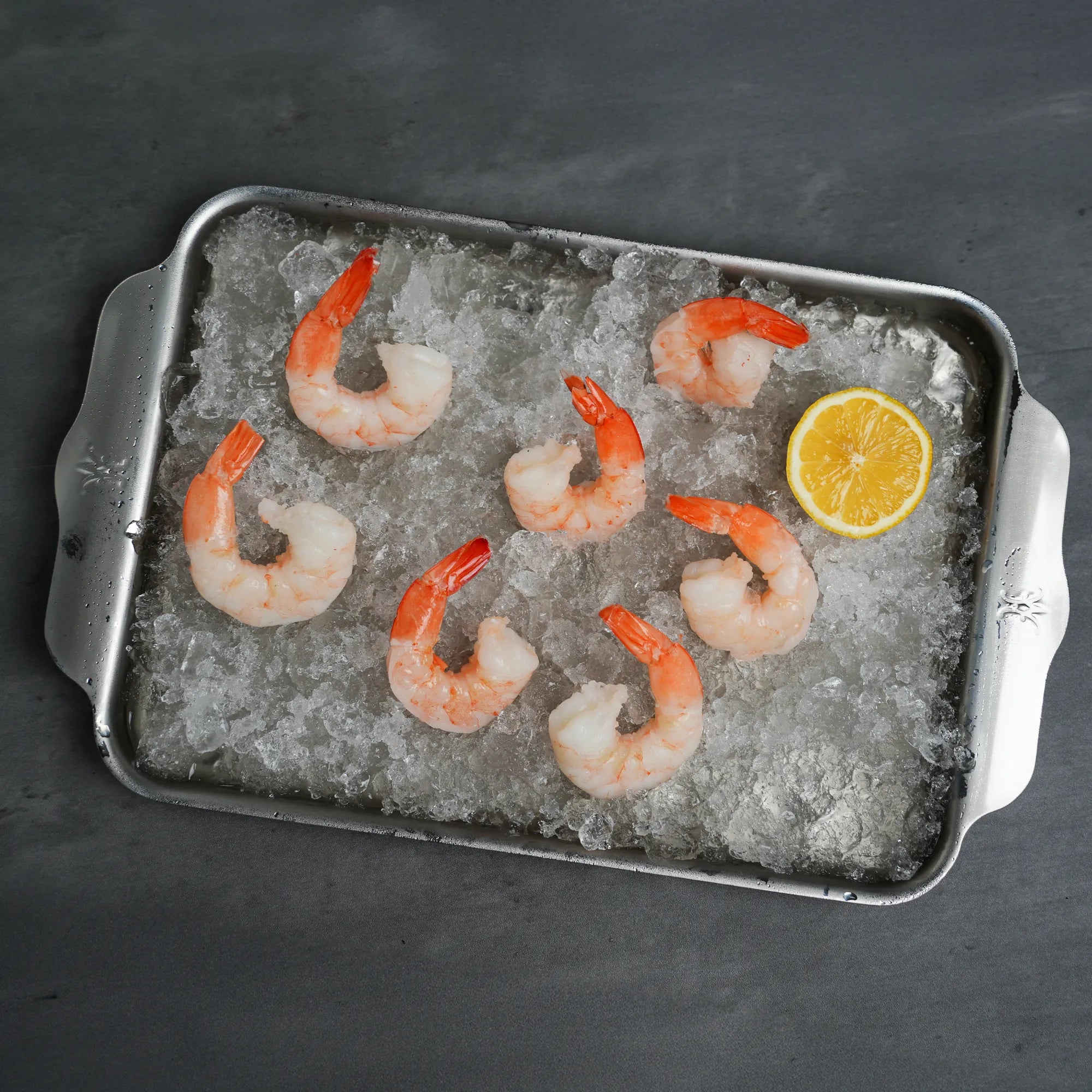 Jumbo Cooked Shrimp arranged on crushed ice with a lemon wedge on a metal tray, showcasing their plumpness and readiness for serving.