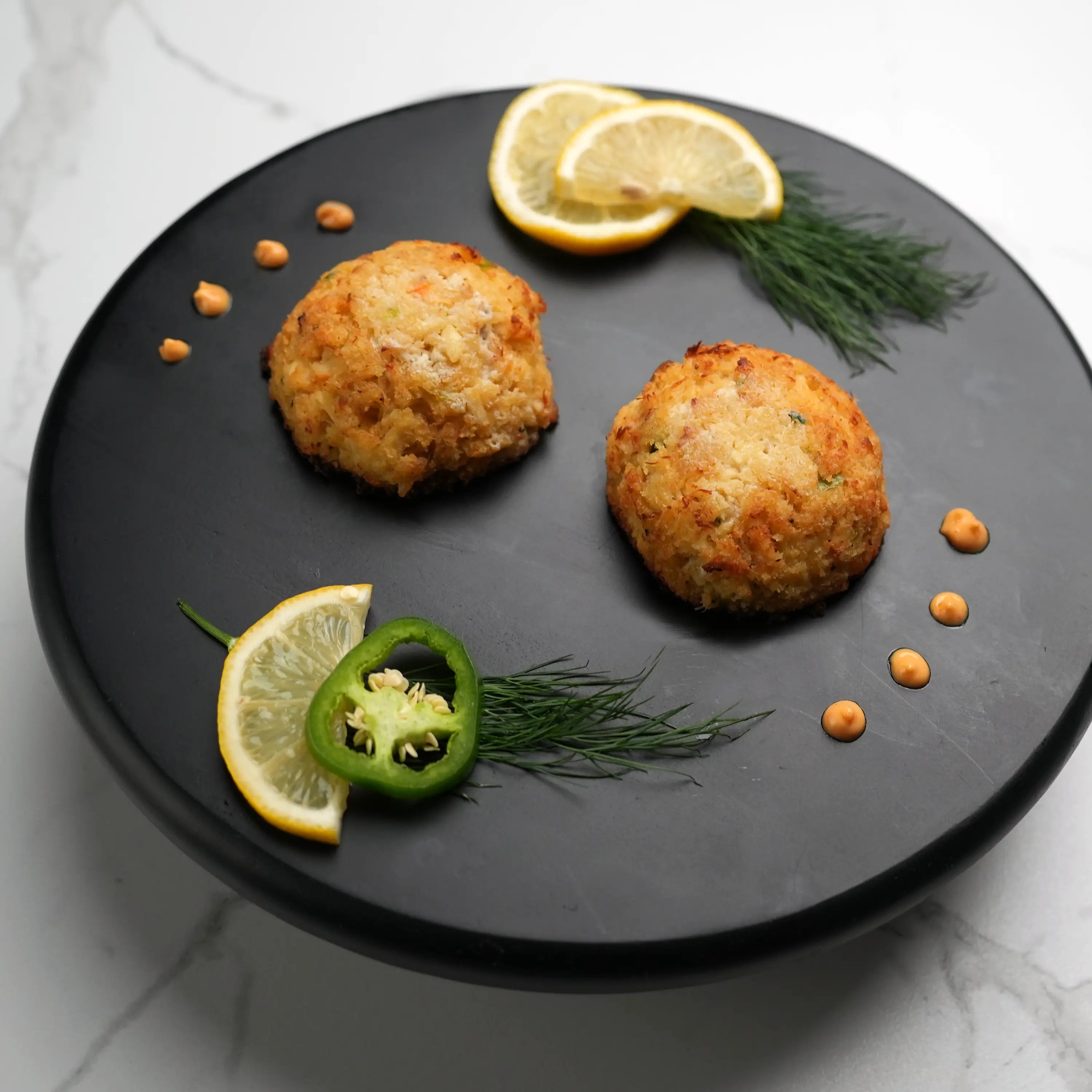 Key West Crab Cakes on a black platter, garnished with lemon wedges, fresh dill, green chili slices, and dots of orange dipping sauce.