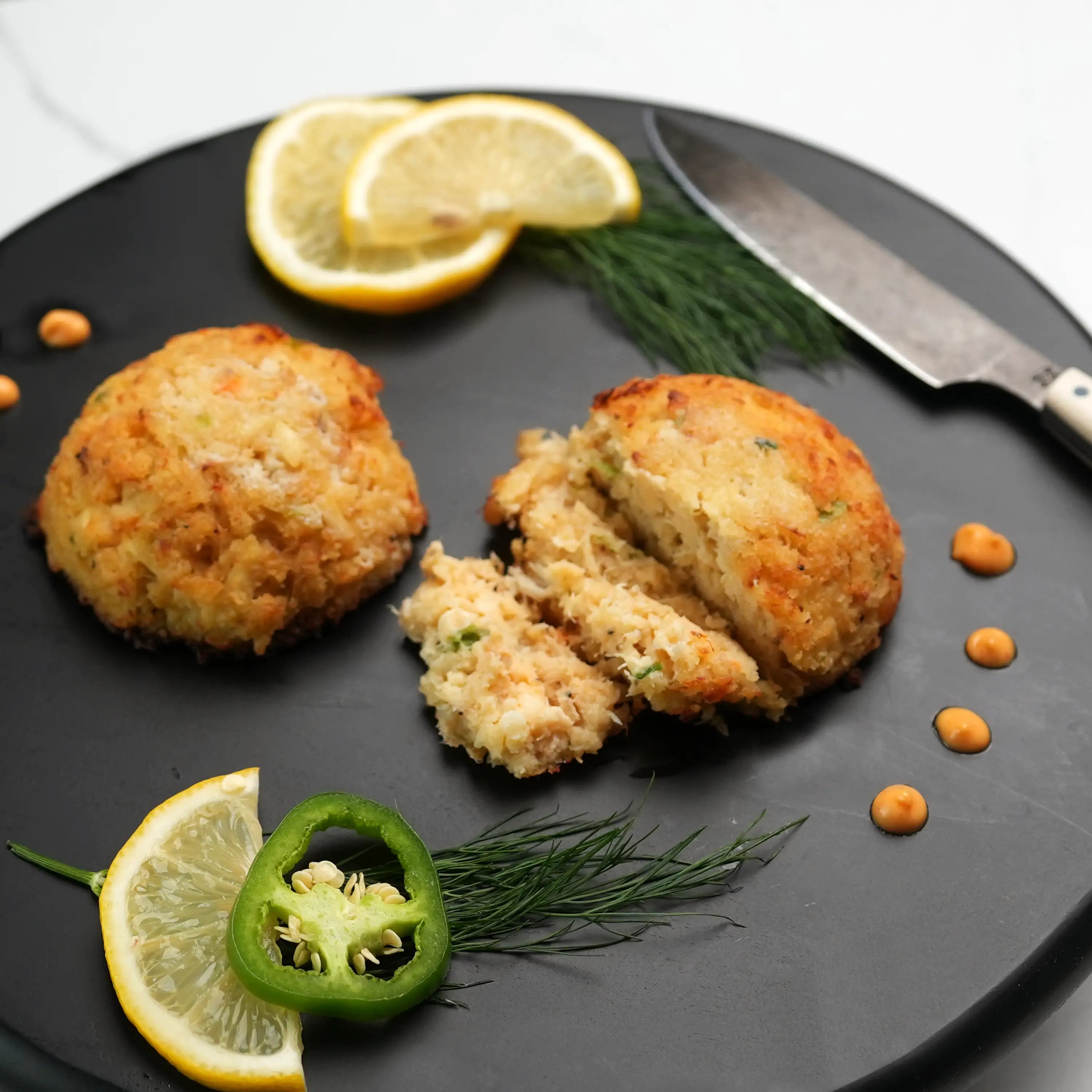Key West Crab Cakes on a black plate, showcasing a flaky interior, garnished with lemon, dill, jalapeño, and orange sauce, with a knife nearby.
