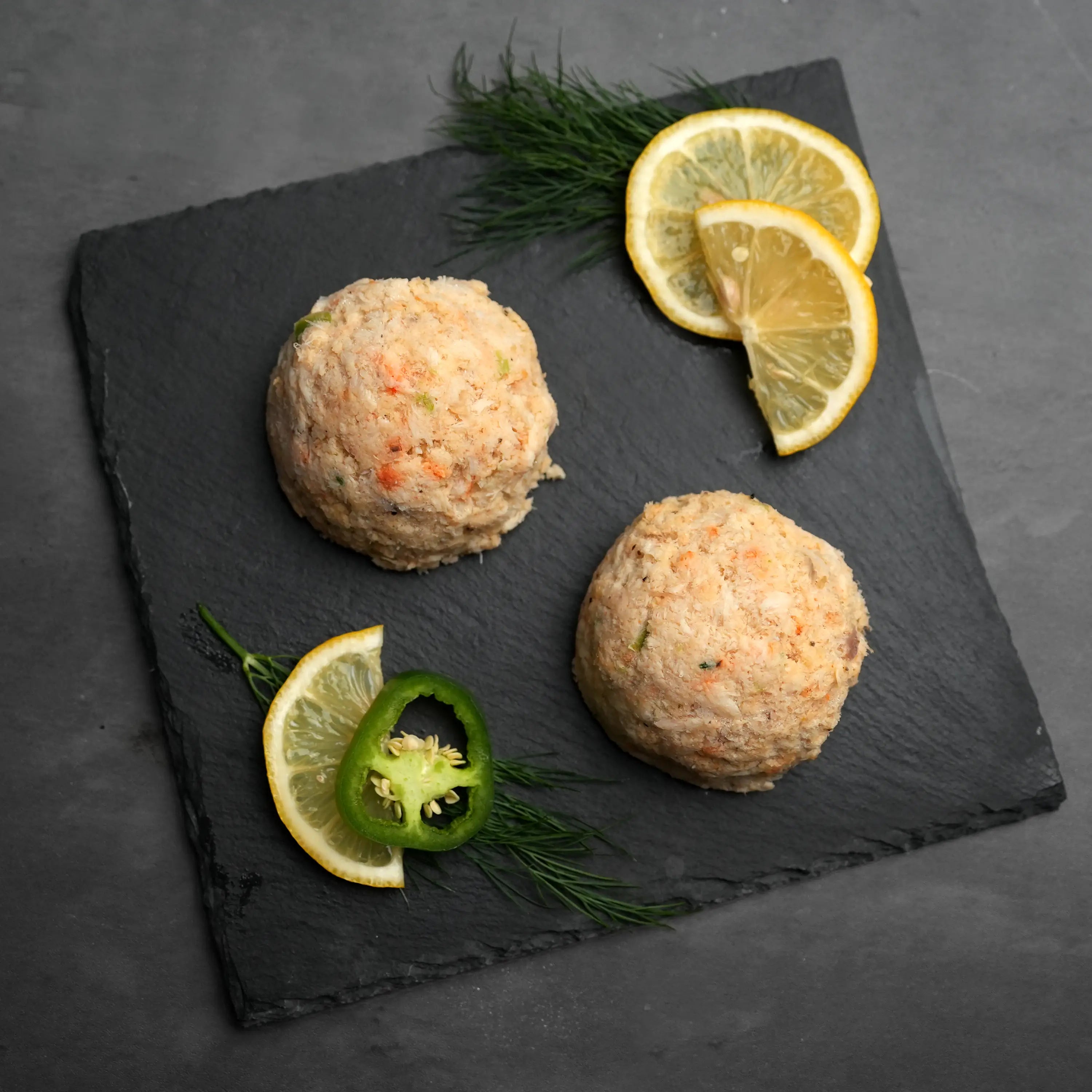 Key West Crab Cakes displayed as three scoops on slate, garnished with dill, lemon wedges, and jalapeño, highlighting their fresh, flavorful seafood composition.