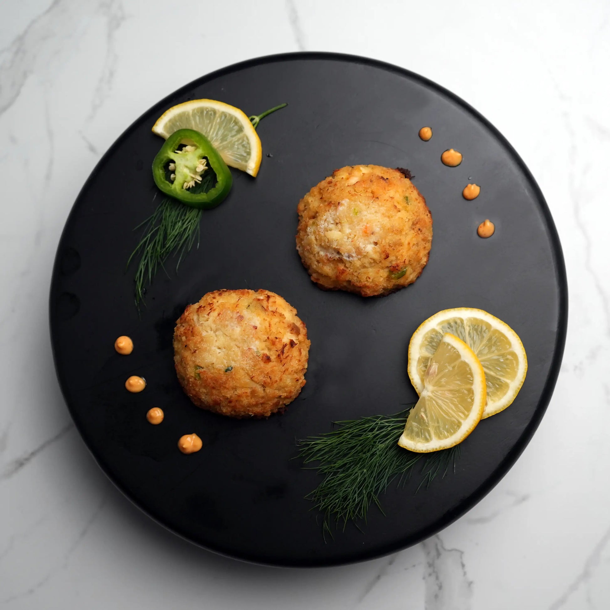 Key West Crab Cakes on a black plate, garnished with dill, lemon slices, and chili rings, accompanied by dots of orange dipping sauce.