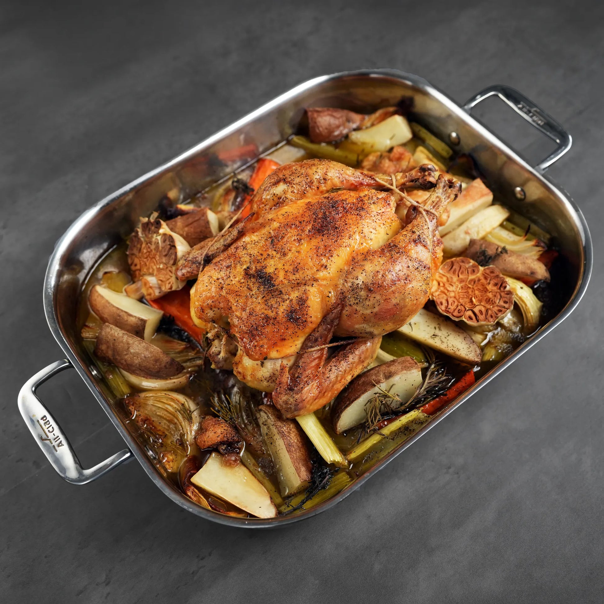 Pasture Raised Heritage Whole Chicken, roasted with cracked pepper, surrounded by roasted vegetables and herbs in a stainless-steel pan, showcasing its superior quality and taste.
