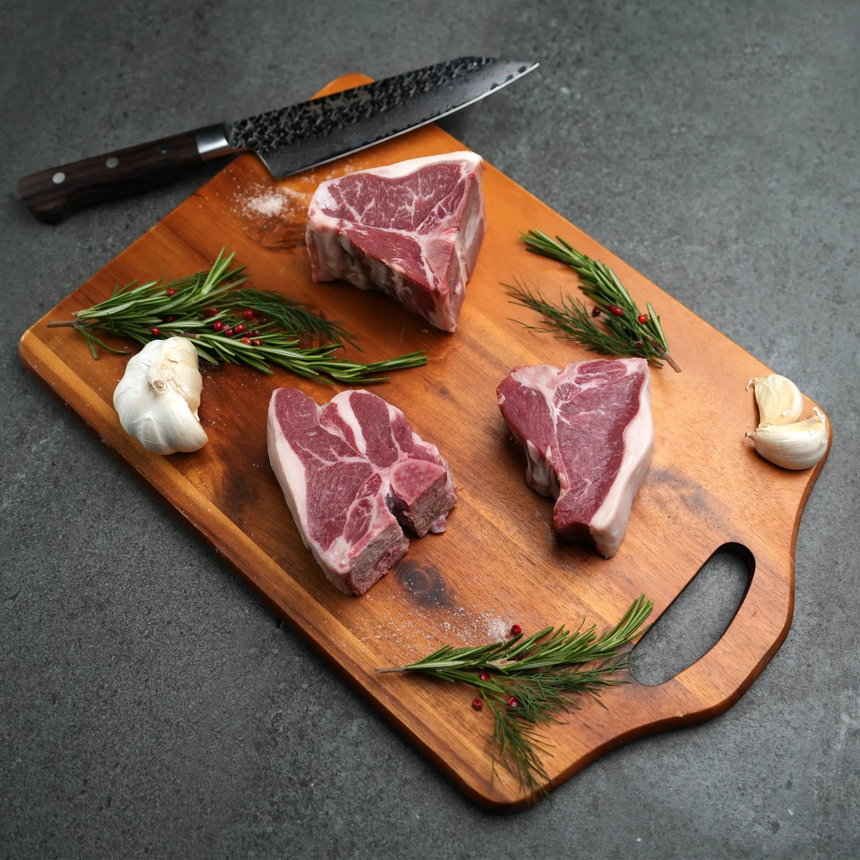 Domestic All Natural Lamb Loin T-Bone Chops on a wooden board with rosemary, garlic, and peppercorns, showcasing their tender, meaty appeal for simple, flavorful cooking.