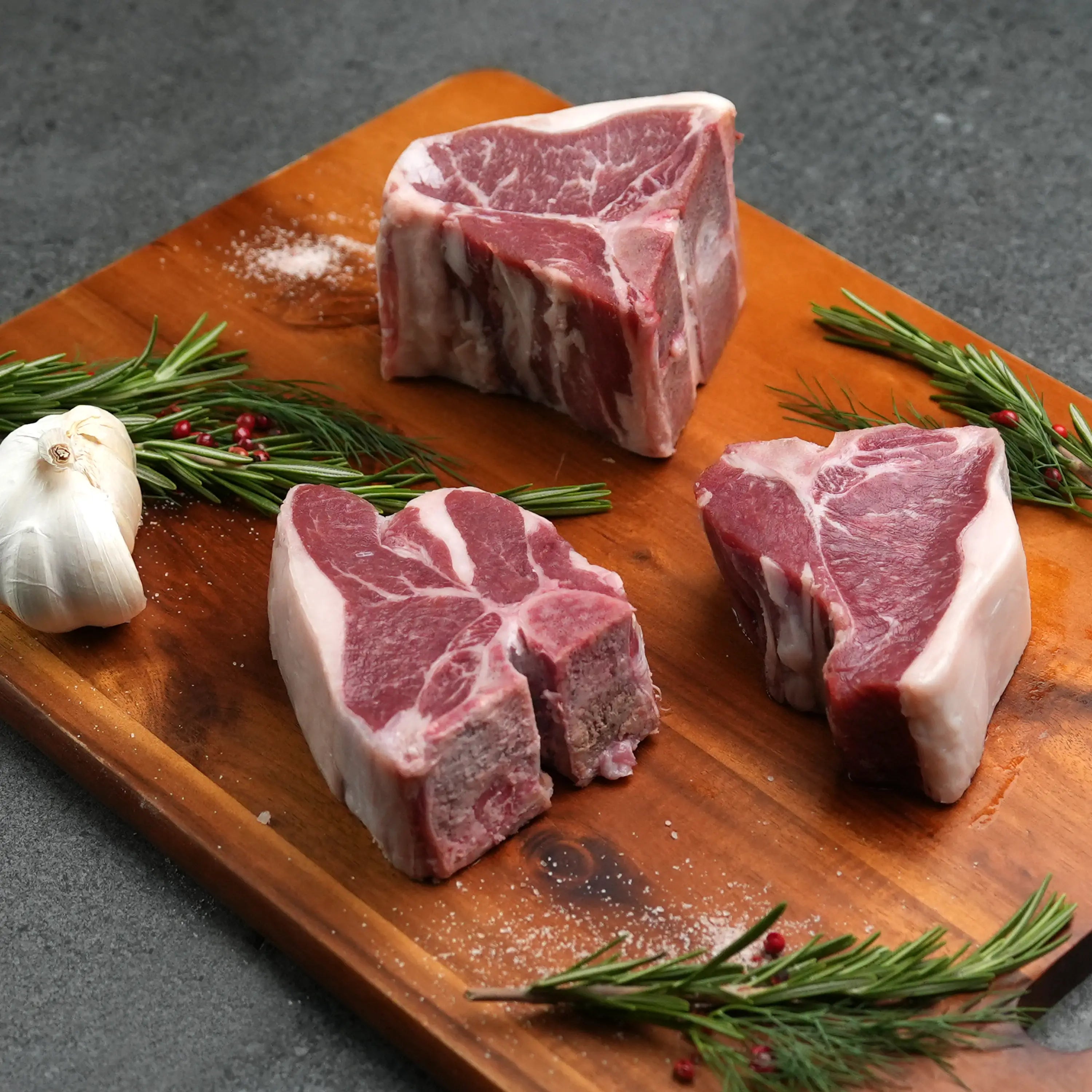 Domestic All Natural Lamb Loin T-Bone Chops on a wooden board with rosemary, garlic, peppercorns, and salt, showcasing their tender, mild flavor ideal for simple preparation.