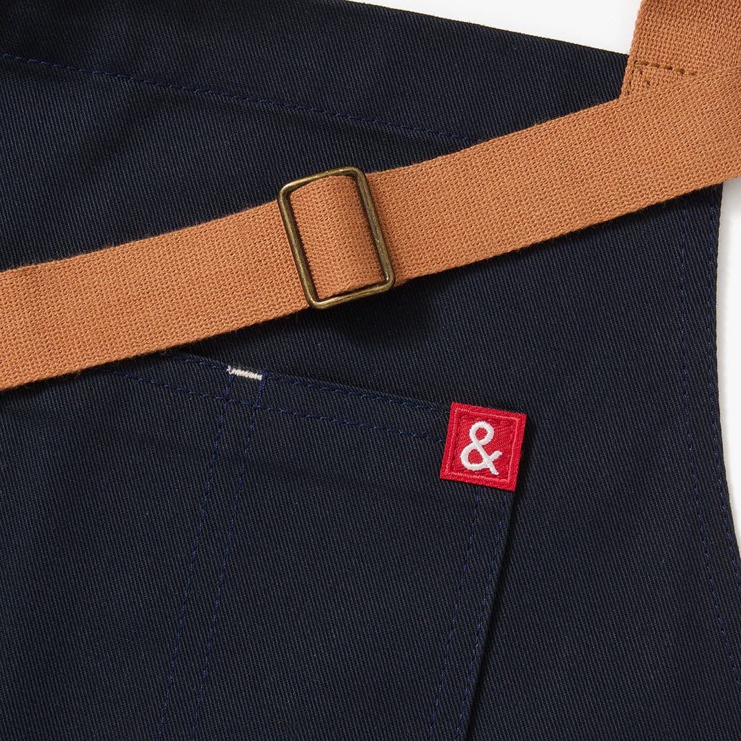 Midnight blue apron detail with tan belt buckle strap and red square logo patch
