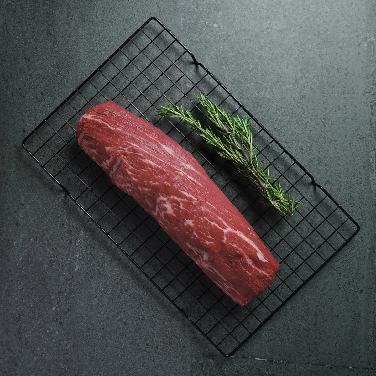 A raw marbled beef roast rests on a black wire cooling rack over a dark gray surface, with a sprig of rosemary placed beside it.