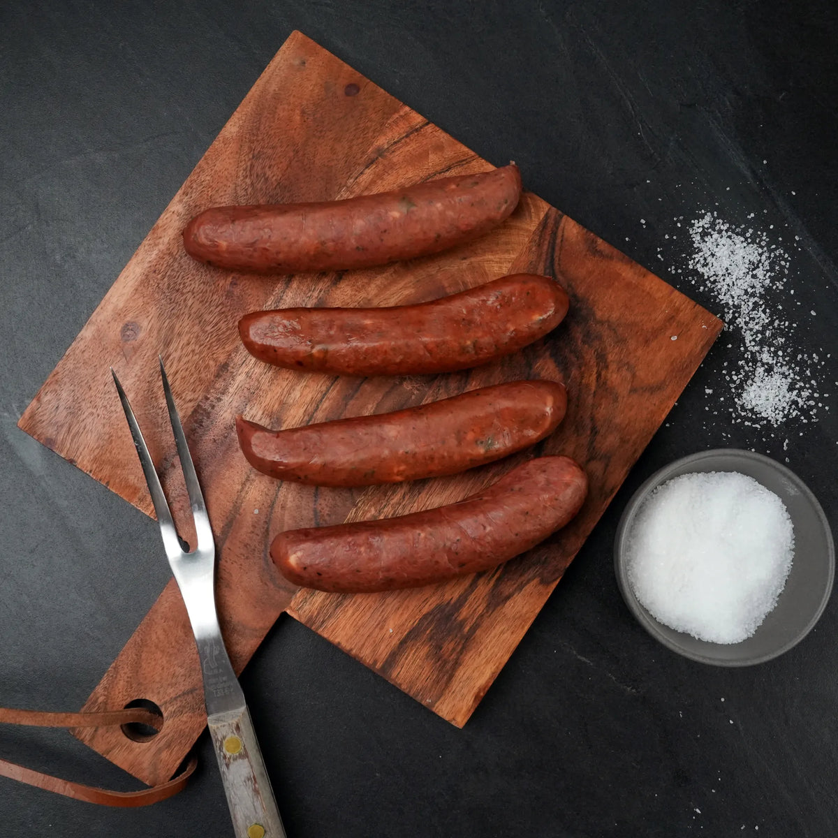 Rosewood Texas Wagyu Jalapeño Queso Sausage links on a wooden board with a meat fork and coarse salt, highlighting their artisanal preparation and gourmet appeal.