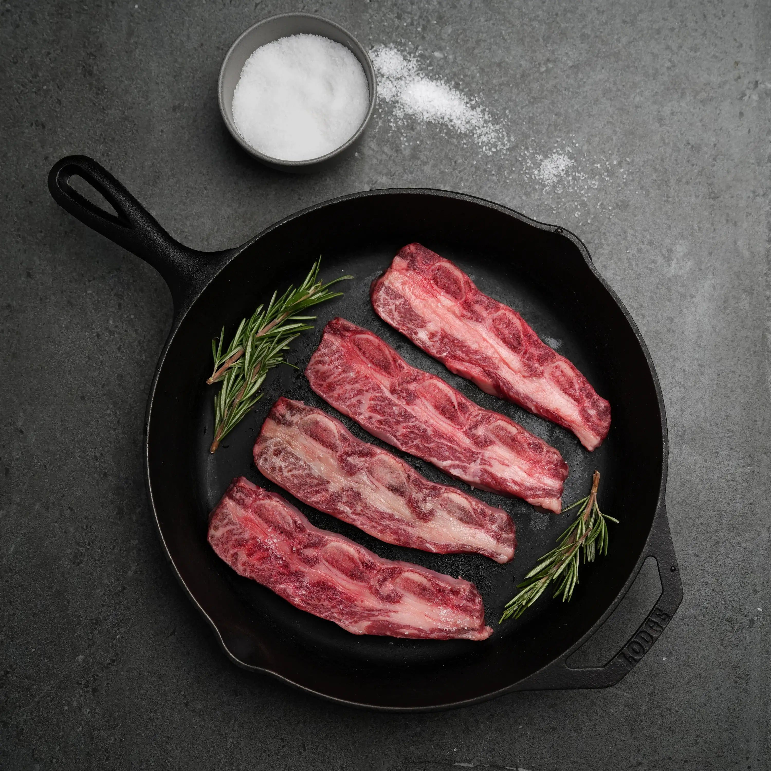 Top-down view of Rosewood Texas Wagyu Flanken Korean Cut Shortribs in a skillet with salt and rosemary, showcasing marbling and preparation for Kalbi.