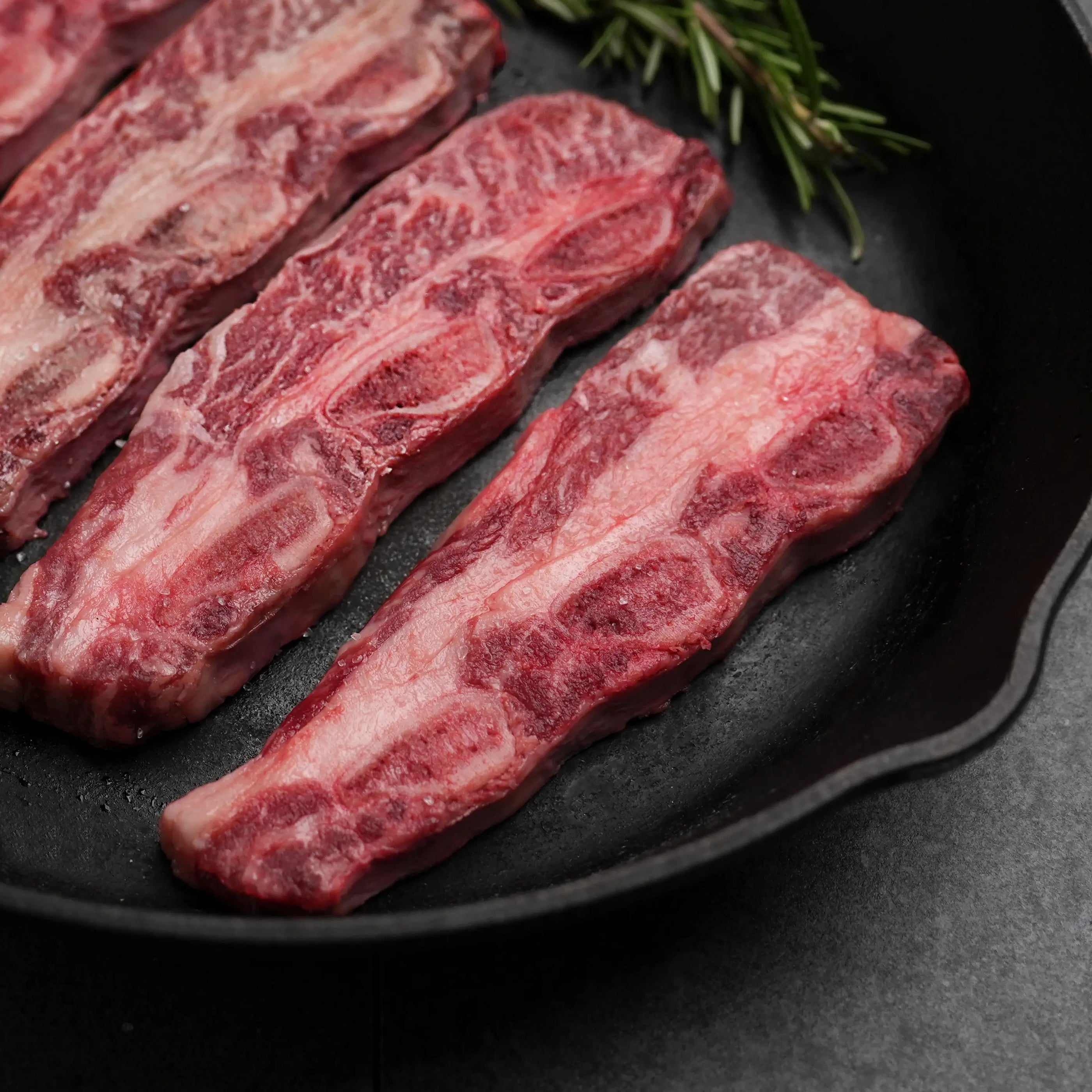 Rosewood Texas Wagyu Flanken Korean Cut Short Rib slices in a skillet, showcasing marbling and a sprig of rosemary, ideal for Korean BBQ.