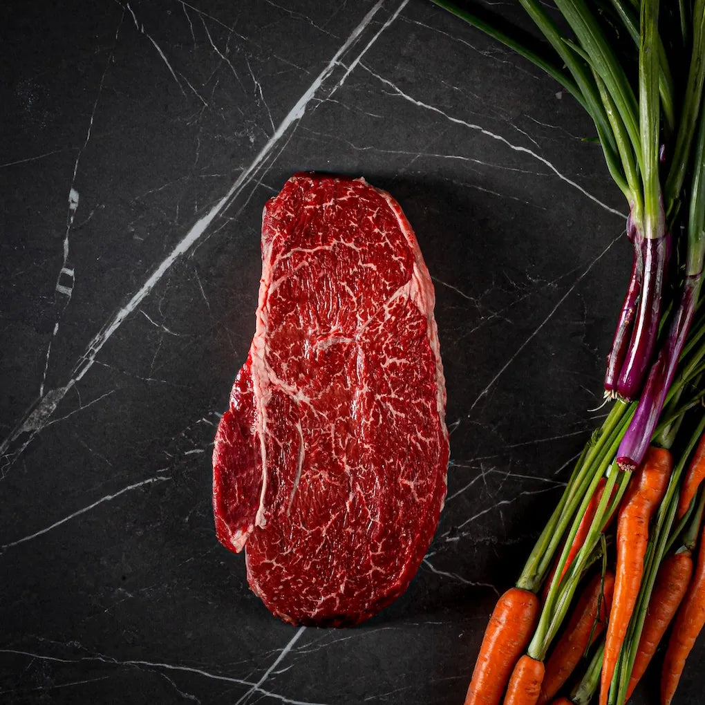 A raw marbled beef steak is photographed from above on a dark marble surface, with carrots and scallions placed along the right side as styling props.