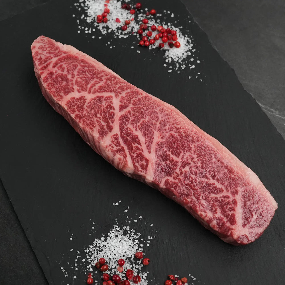 Stone Axe Australian Coulotte Cap Steak, a thick-cut, marbled raw beef piece on slate with coarse sea salt and scattered pink peppercorns.