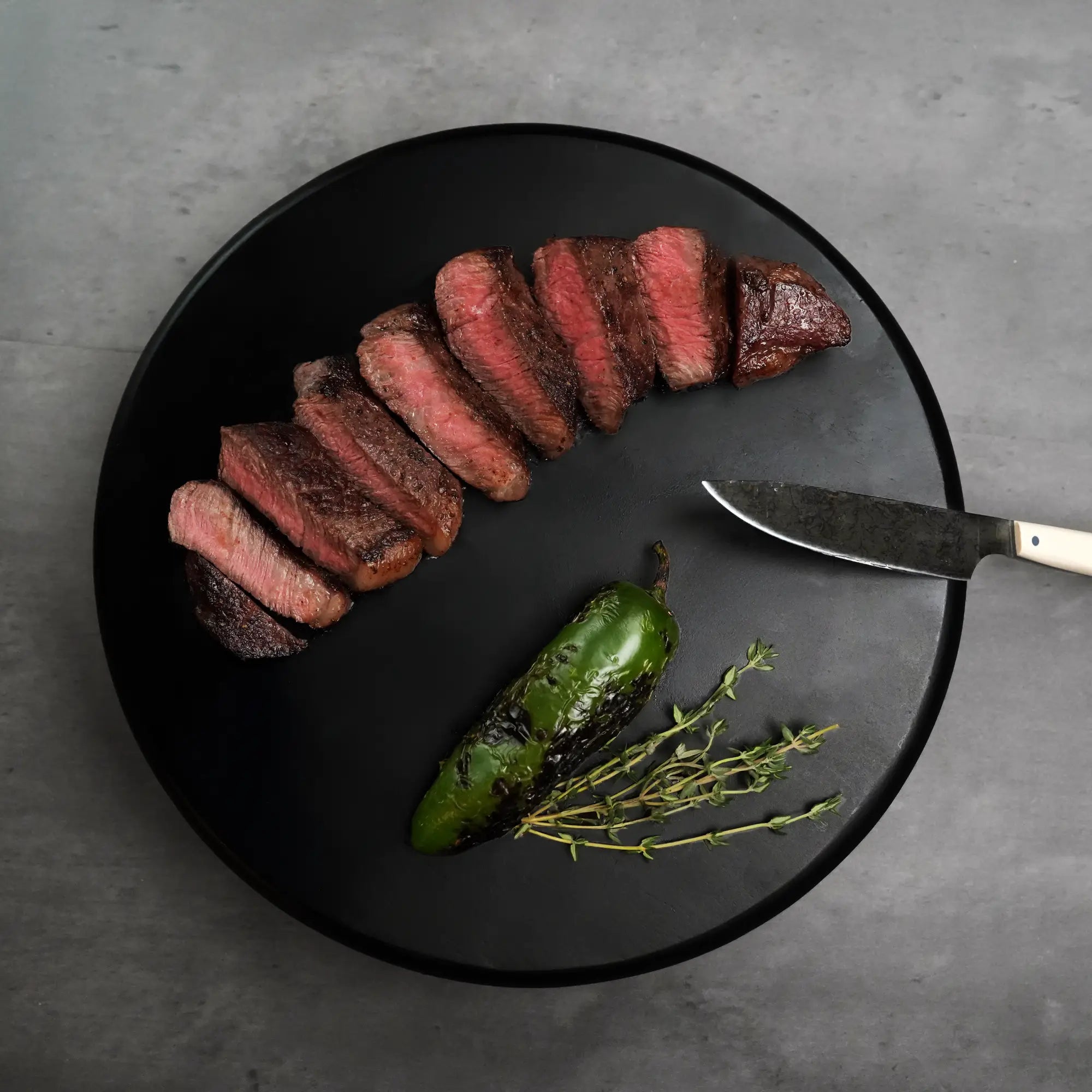 Stone Axe Australian Coulotte Cap Steak, medium-rare slices on a dark plate with charred green pepper, fresh thyme, and a carving knife alongside.