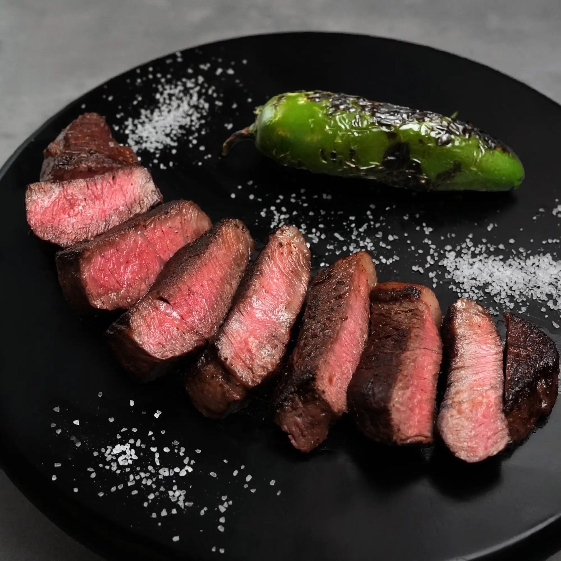 Stone Axe Australian Coulotte Cap Steak slices on a plate with charred green chili, sprinkled with coarse salt flakes.