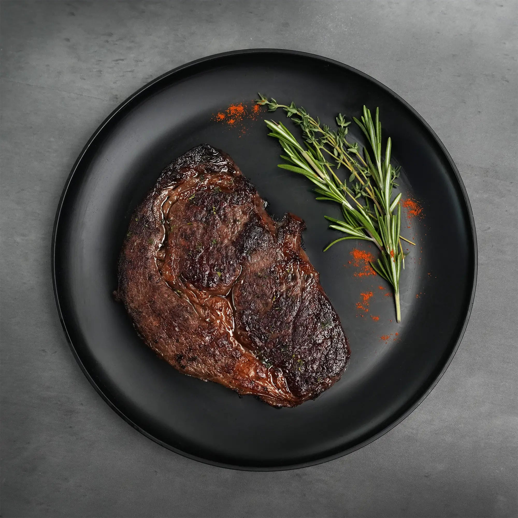 Stone Axe Australian Wagyu Boneless Ribeye Steak on a black plate, seared and glazed, garnished with rosemary, thyme, and red spice on a dark tabletop.