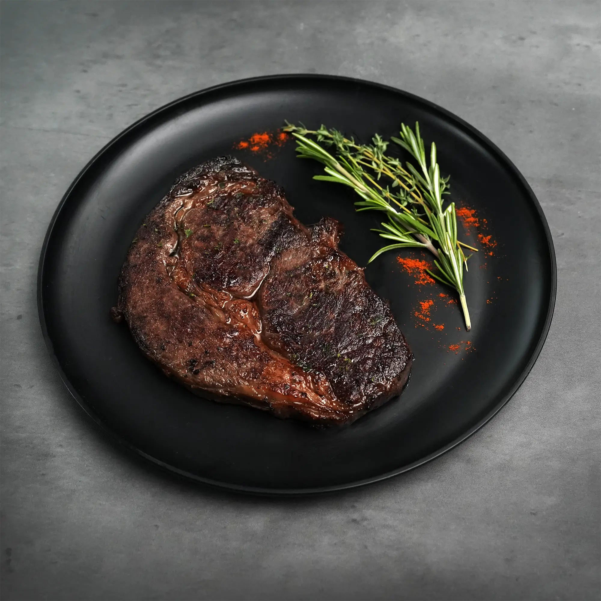 Stone Axe Australian Wagyu Boneless Ribeye Steak on a black plate, garnished with rosemary, thyme, and red spice, highlighting its premium quality and flavor.