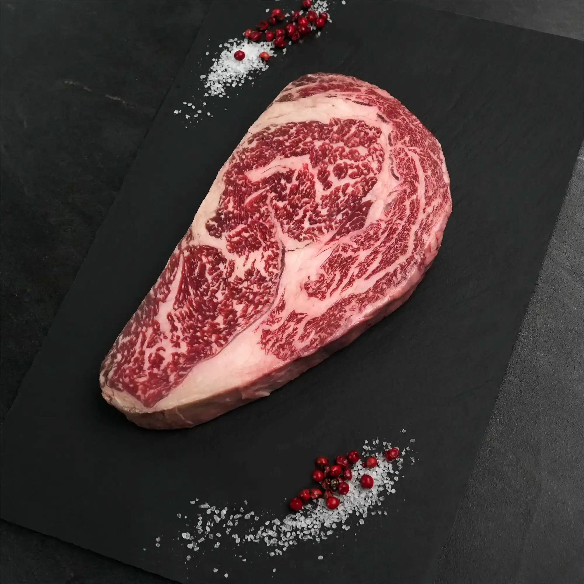 Stone Axe Australian Wagyu Boneless Ribeye Steak on slate, showcasing rich marbling. Surrounded by sea salt and pink peppercorns, ideal for gourmet preparation.