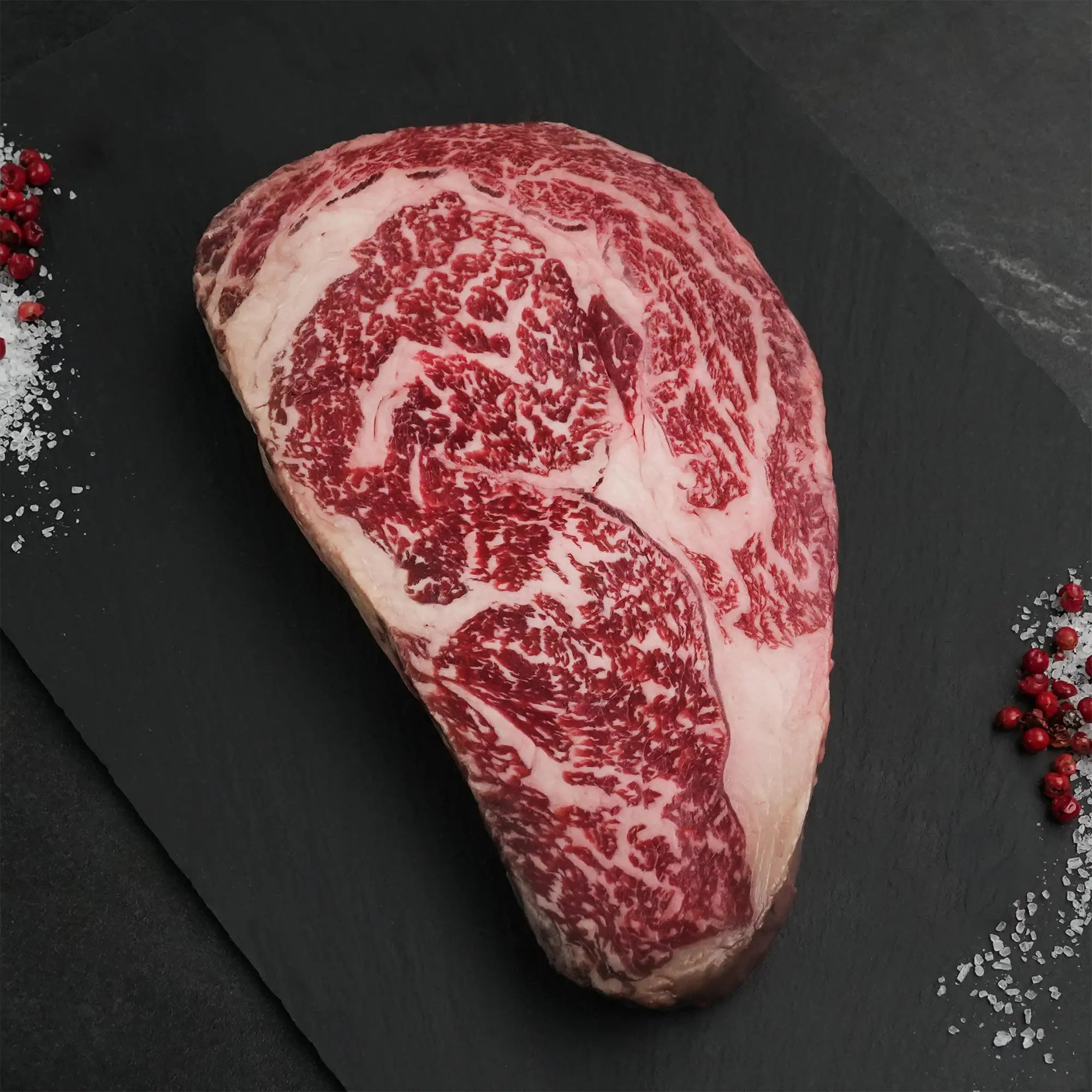Stone Axe Australian Wagyu Boneless Ribeye Steak on slate with marbled texture, coarse sea salt, and red peppercorns scattered beside it.