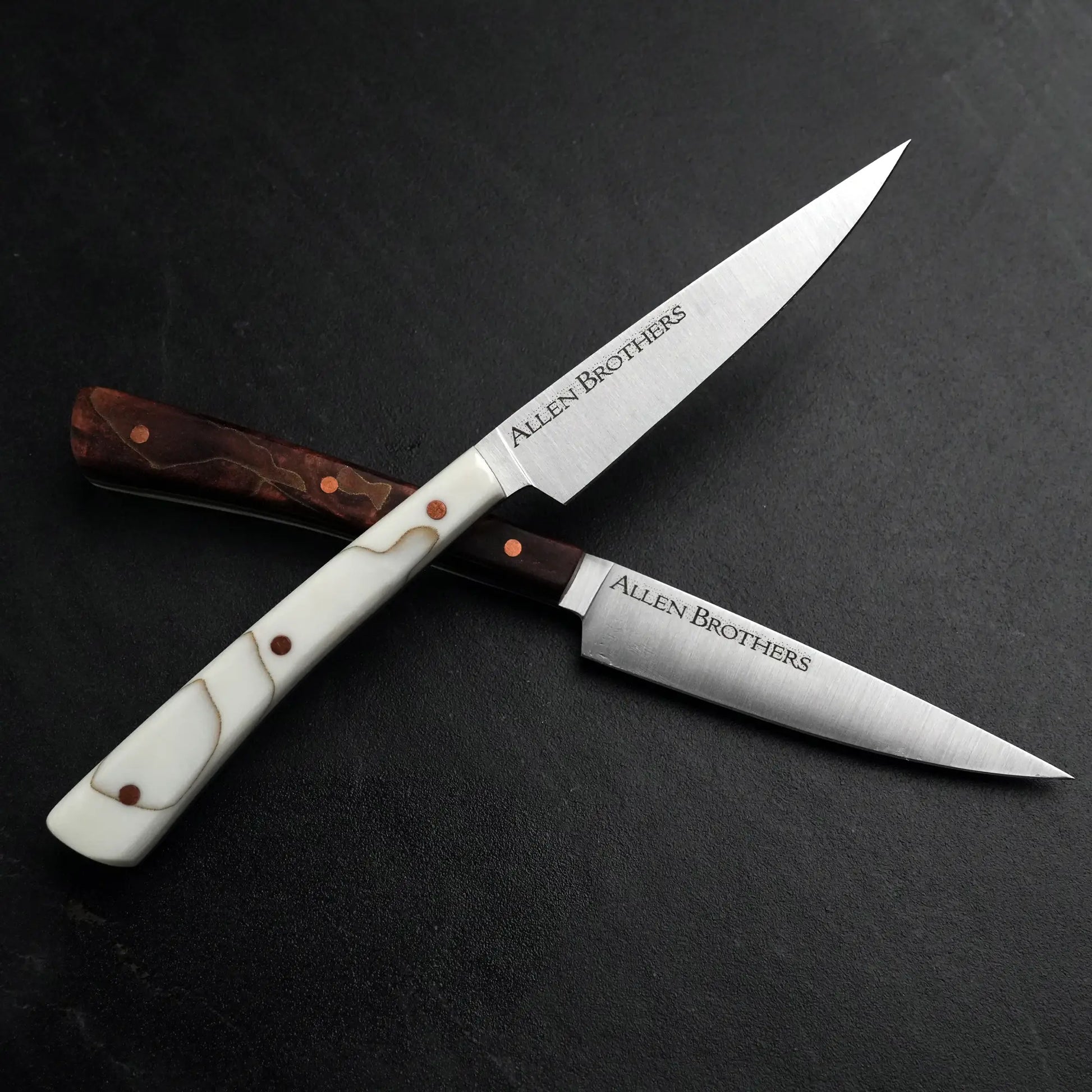 TH Blades Handcrafted Duality Paring Knife Set: Two crossed knives with contrasting handles and copper rivets, etched with “Allen Brothers,” on a dark surface.