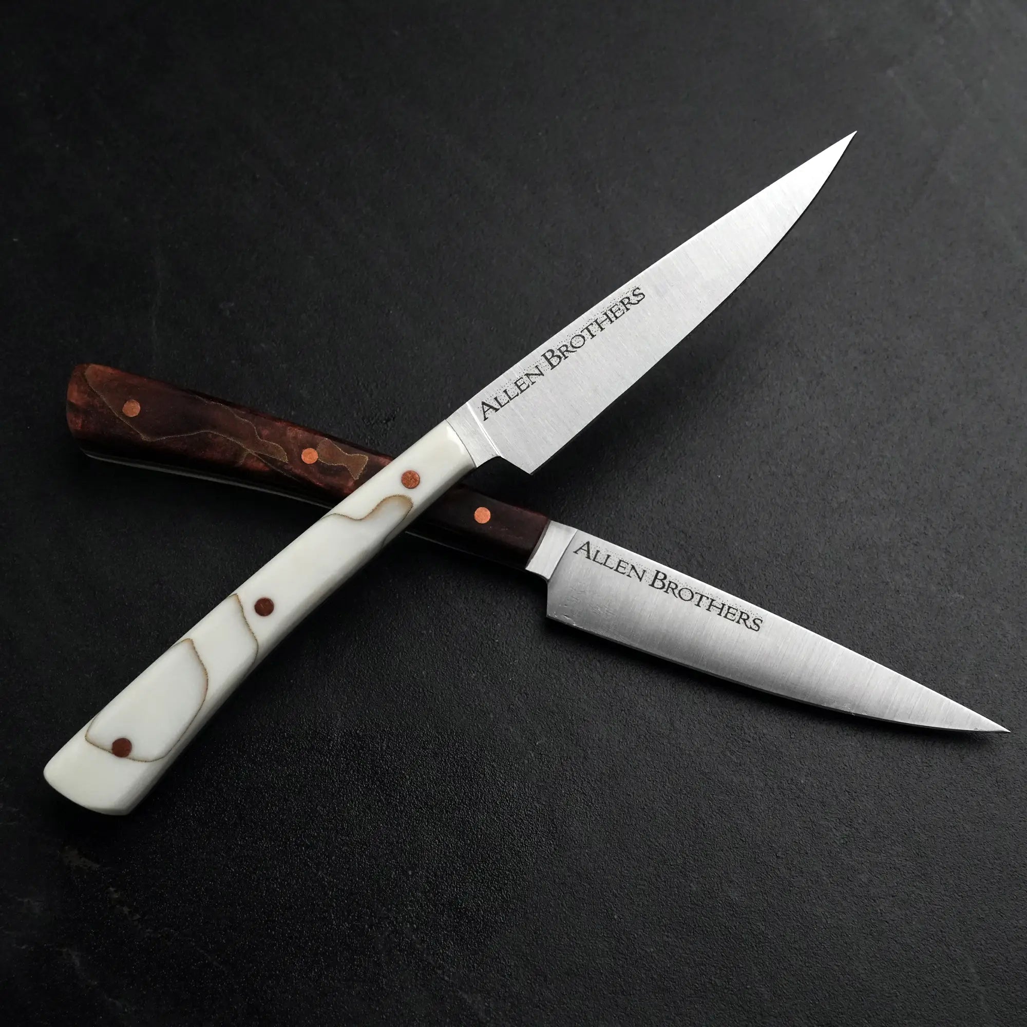 TH Blades Handcrafted Duality Paring Knife Set: Two crossed knives with contrasting handles and copper rivets, etched with “Allen Brothers,” on a dark surface.