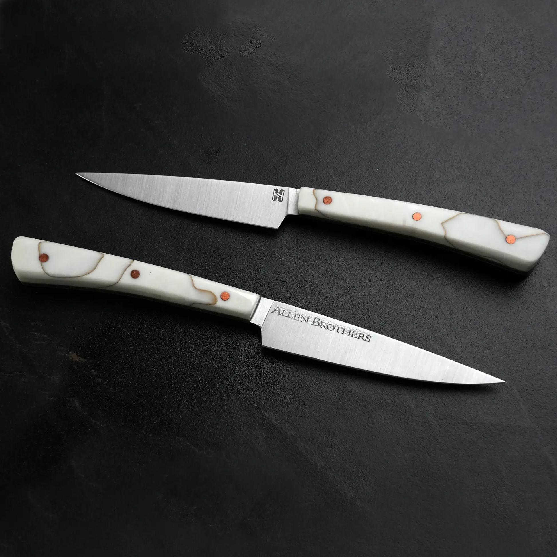 TH Blades Handcrafted Duality Paring Knife Set on dark countertop, featuring two slender knives with stainless-steel blades, white handles, and copper rivets, designed by Todd Harrington.