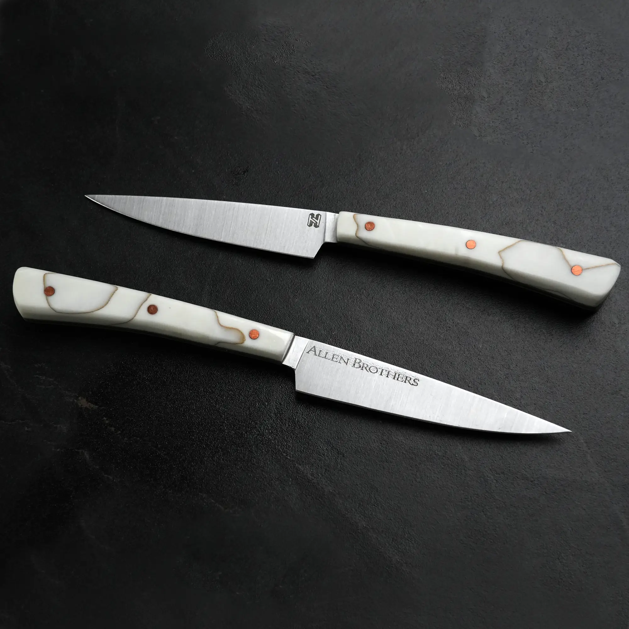 TH Blades Handcrafted Duality Paring Knife Set on dark countertop, featuring two slender knives with stainless-steel blades, white handles, and copper rivets, designed by Todd Harrington.