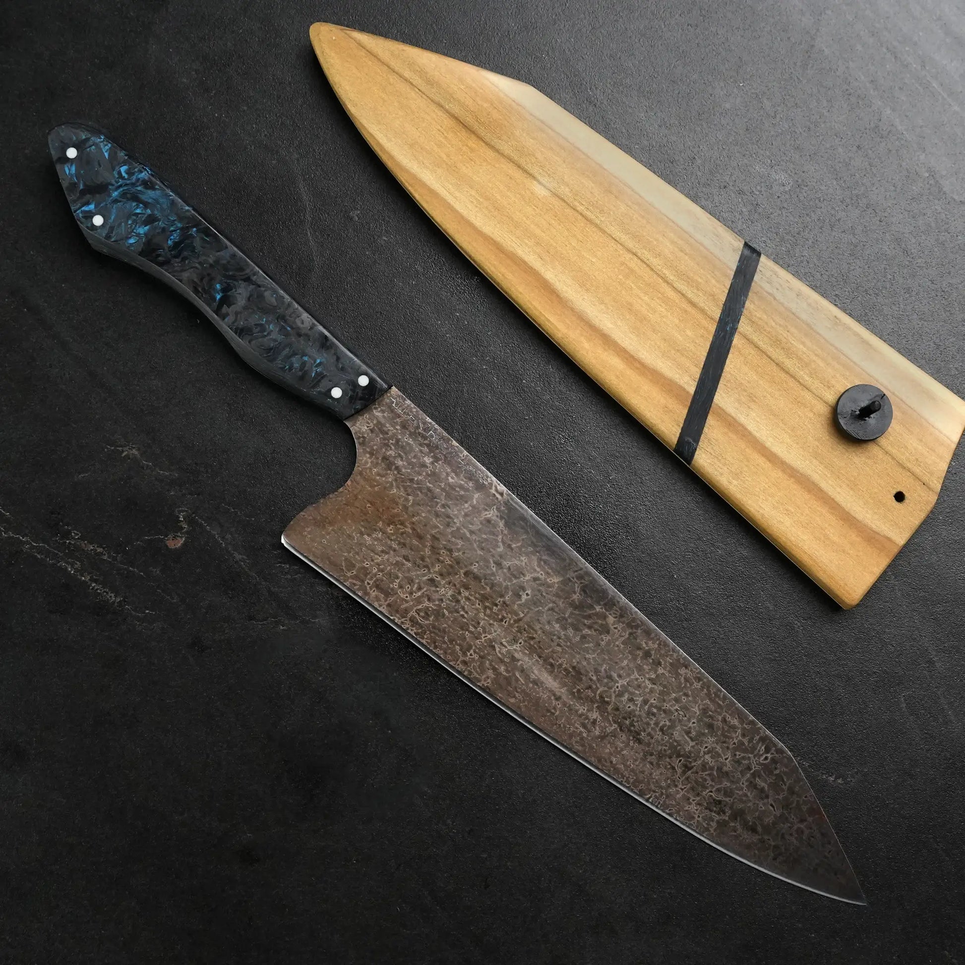 TH Blades Handcrafted 8 Chef Knife with hammered high carbon steel blade and black-blue swirled resin handle, alongside a wooden sheath with a black fastener.