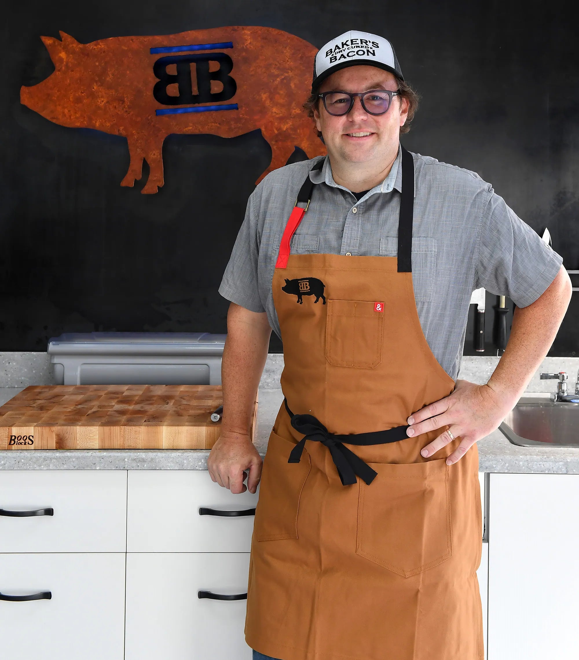 Man wearing a brown apron and a cap with a logo, standing in a kitchen.