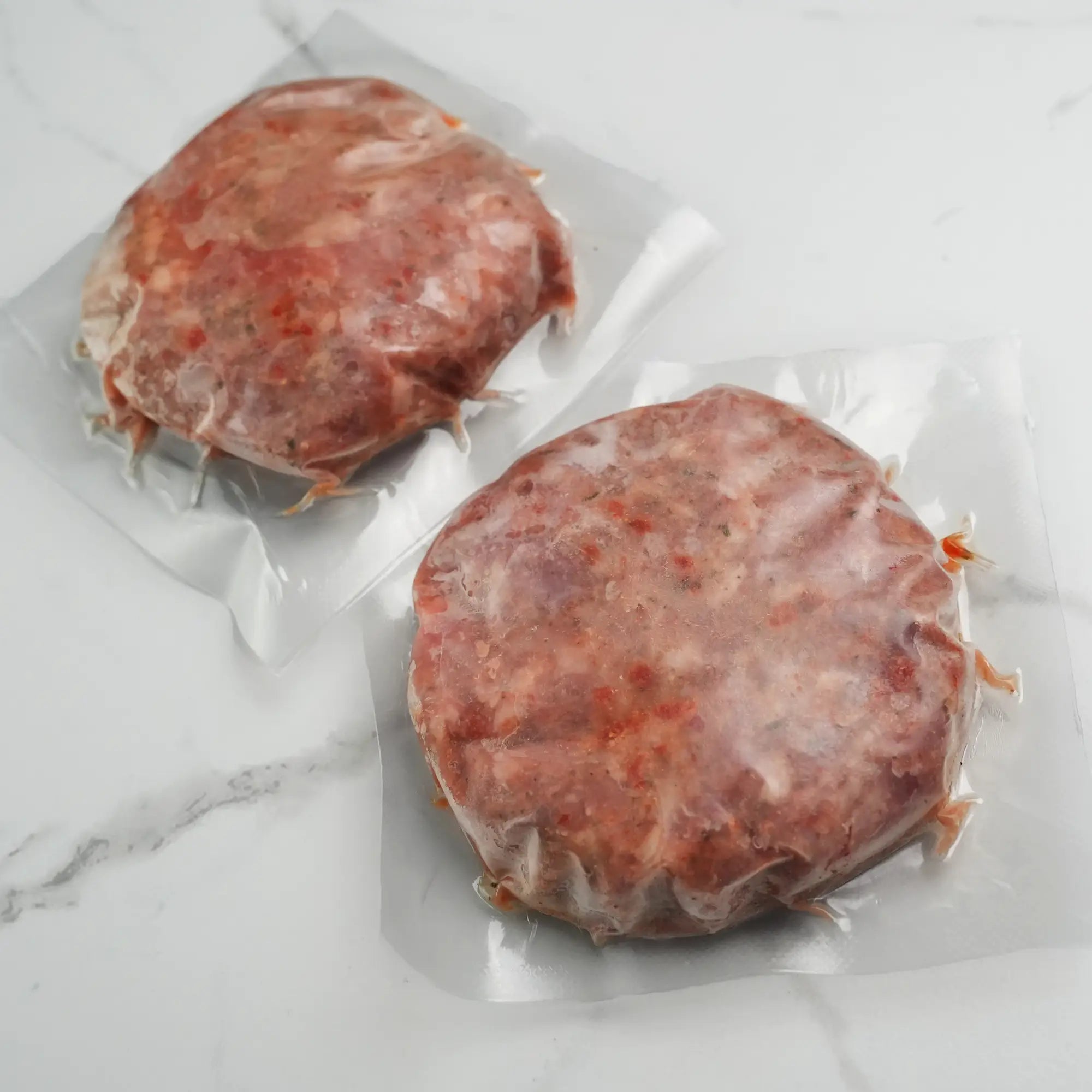 Two vacuum-sealed turkey burgers on a marble countertop, shaped and seasoned with pepper, onion, and soy sauce, ready for grilling.