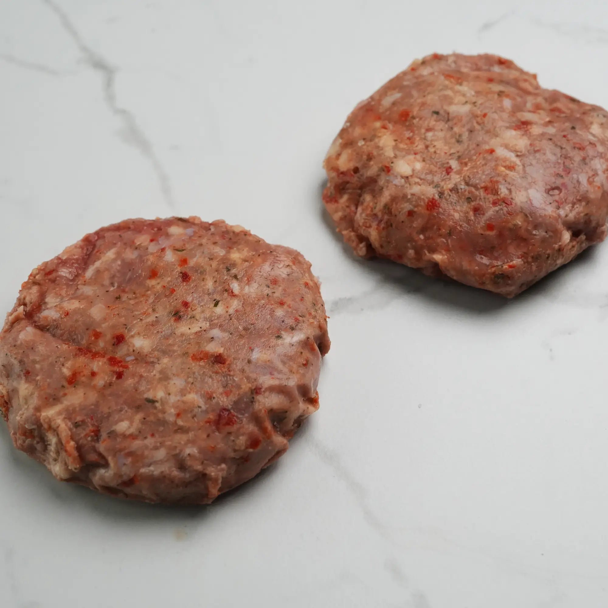 Two uncooked, seasoned turkey burger patties on a white surface, showcasing flecks of pepper and onion, ready for grilling.