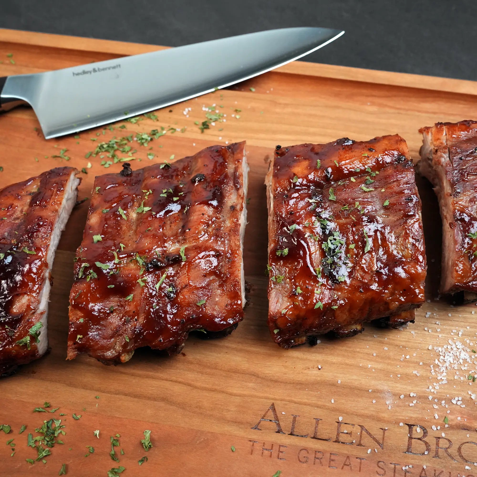 Heritage Duroc Pork Peeled Baby Back Ribs sliced on a wooden board, topped with sauce, coarse salt, and herbs, with a chef’s knife in the background.