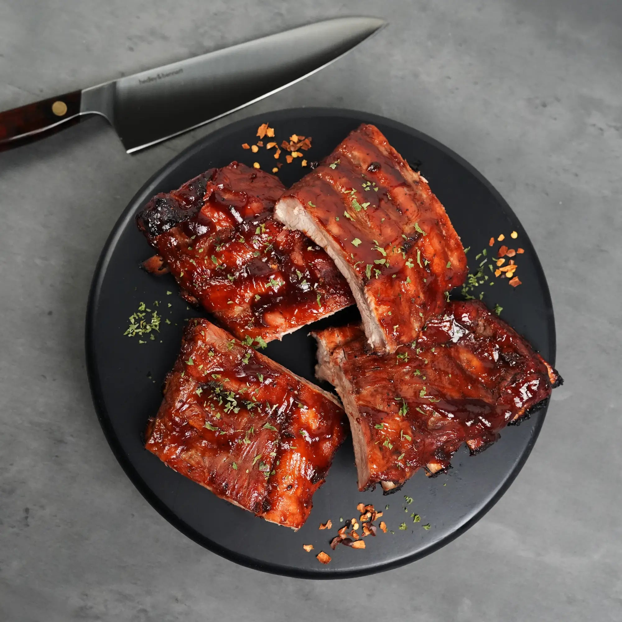 Heritage Duroc Pork Peeled Baby Back Ribs, glazed and garnished with herbs and red-pepper flakes, sliced on a black plate beside a chef's knife.