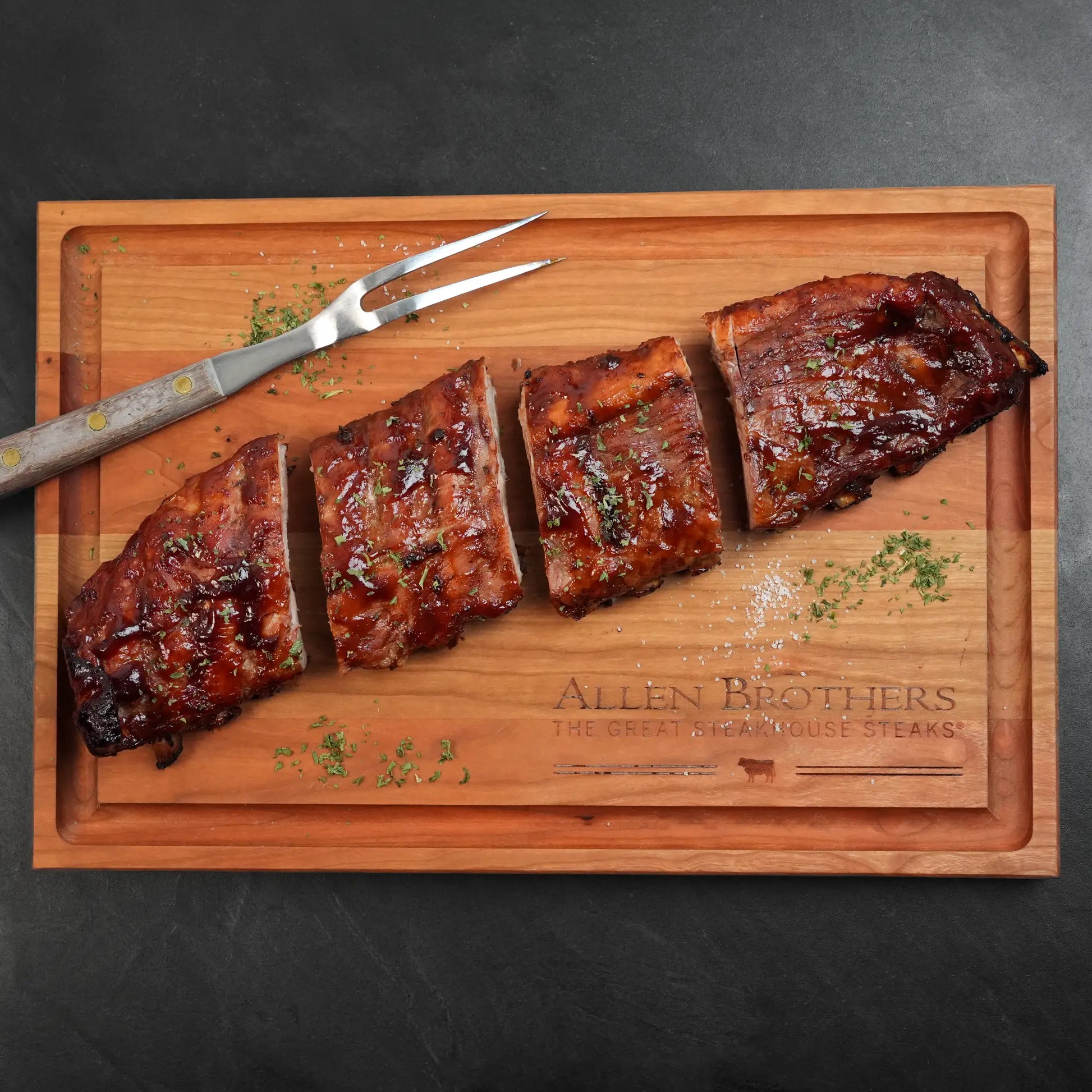 Heritage Duroc Pork Peeled Baby Back Ribs on a wooden carving board, partially sliced and glazed, sprinkled with salt and herbs, accompanied by a carving fork.
