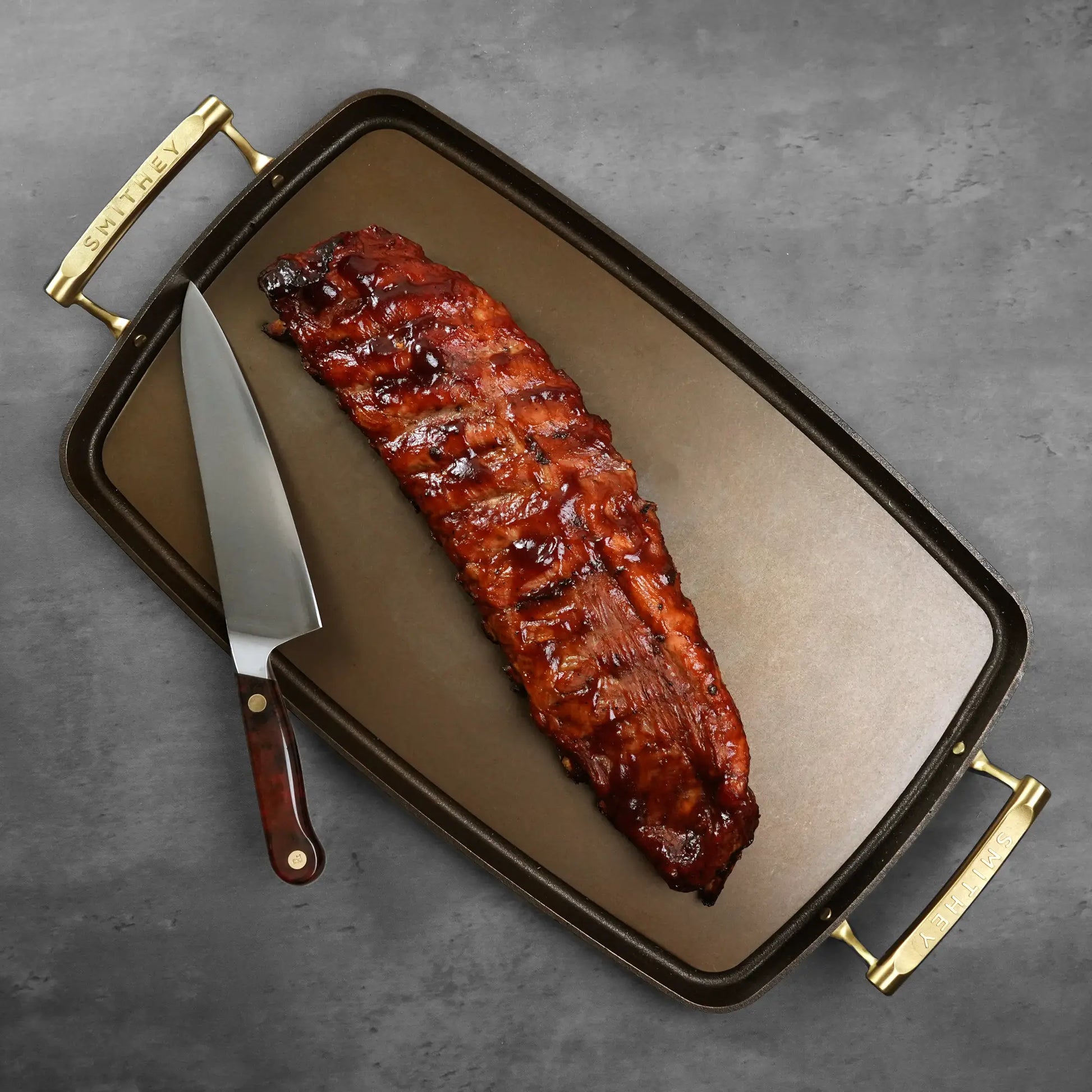 Heritage Duroc Pork Peeled Baby Back Ribs in a Smithey cast-iron pan with brass handles, accompanied by a large carving knife.