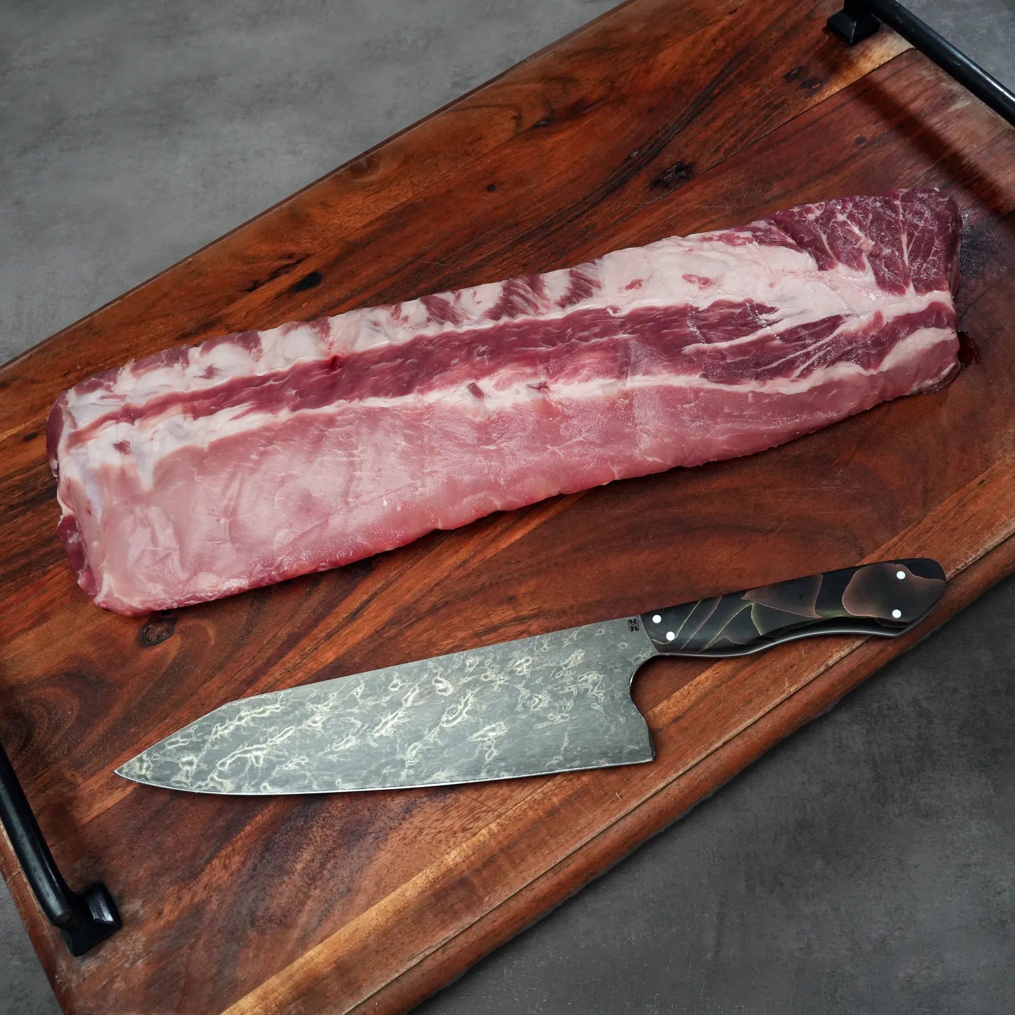 Heritage Duroc Pork Peeled Baby Back Ribs on a wooden board with a Damascus-style knife, ready for seasoning and cooking.