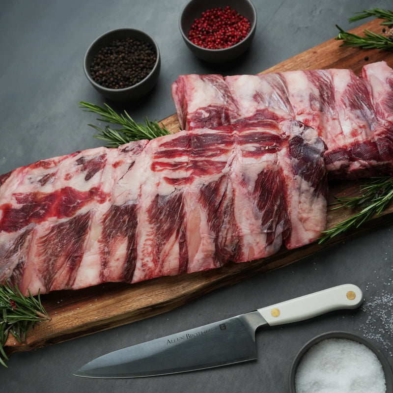 Rosewood Texas Wagyu Beef Back Rib on a board with rosemary, peppercorns, sea salt, and an Allen Brothers knife, highlighting its marbling and tenderness.