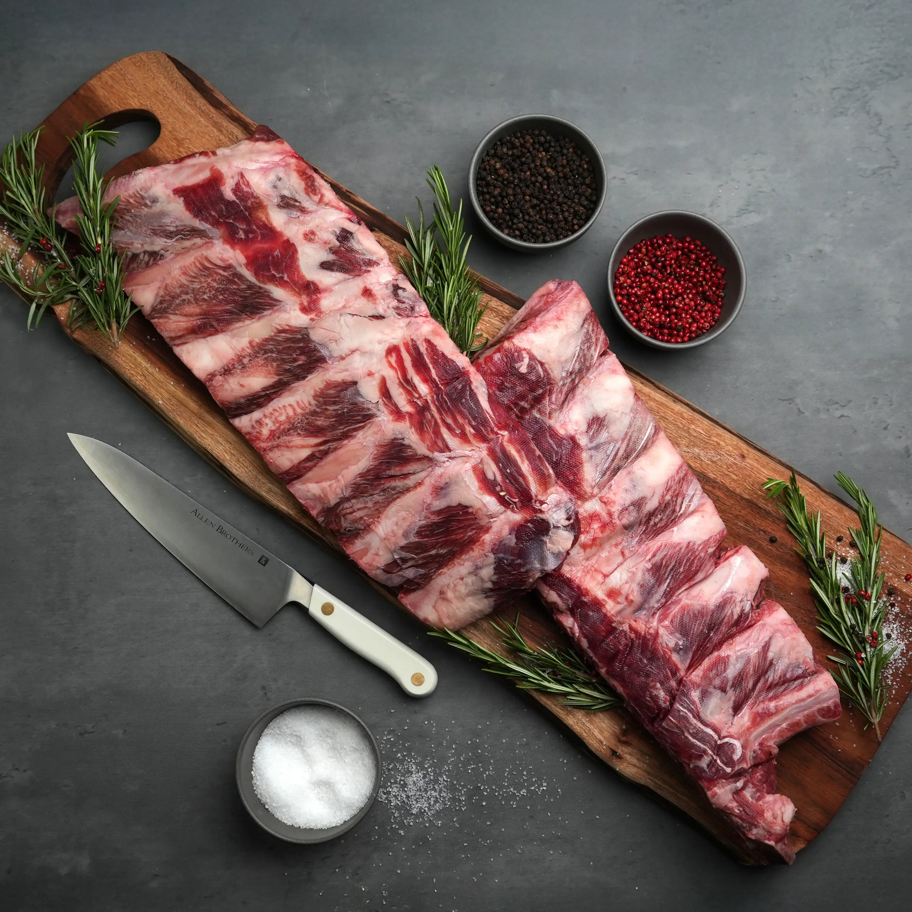 Rosewood Texas Wagyu Beef Back Rib on wooden board with rosemary, salt, peppercorns; chef’s knife nearby. Perfectly marbled for grilling or smoking.