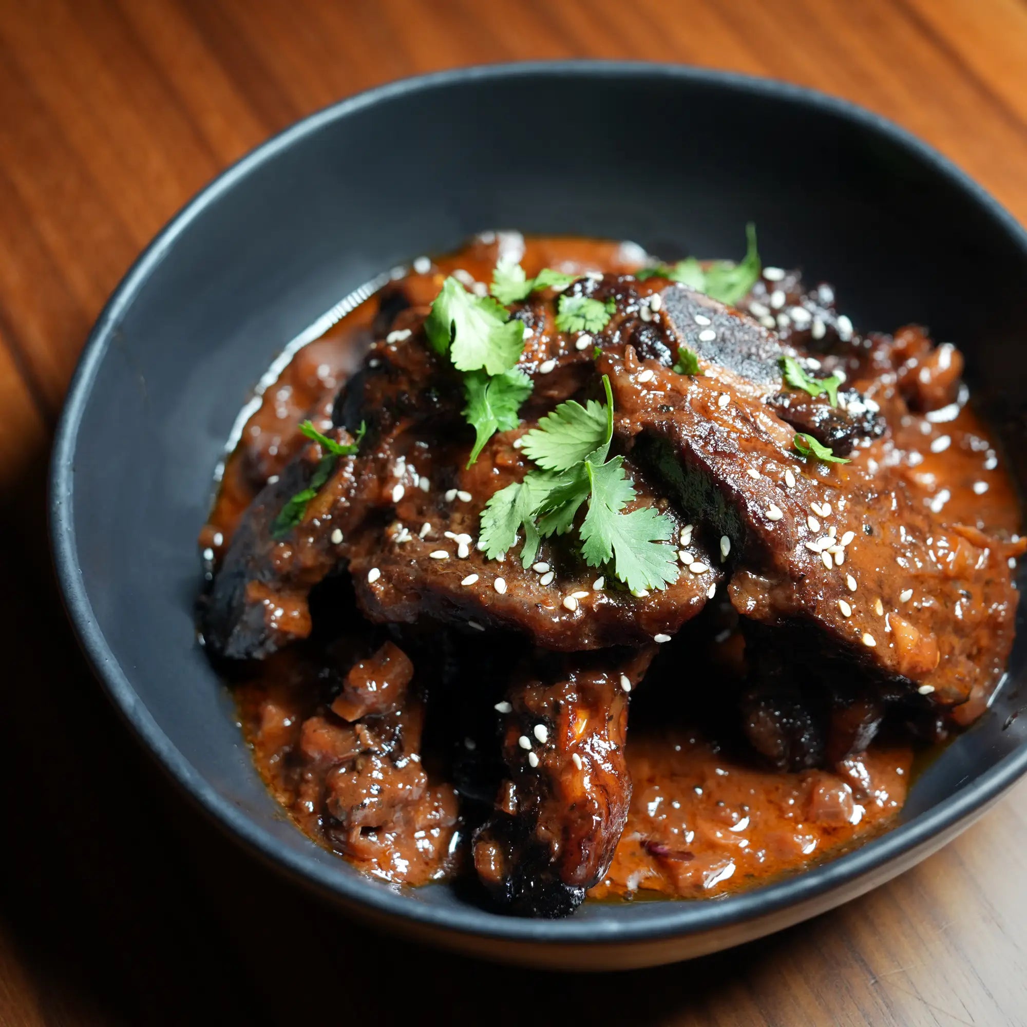 Rosewood Texas Wagyu Flanken Korean Cut Shortrib, char-grilled with reddish-brown sauce, garnished with cilantro and sesame seeds in a dark bowl.