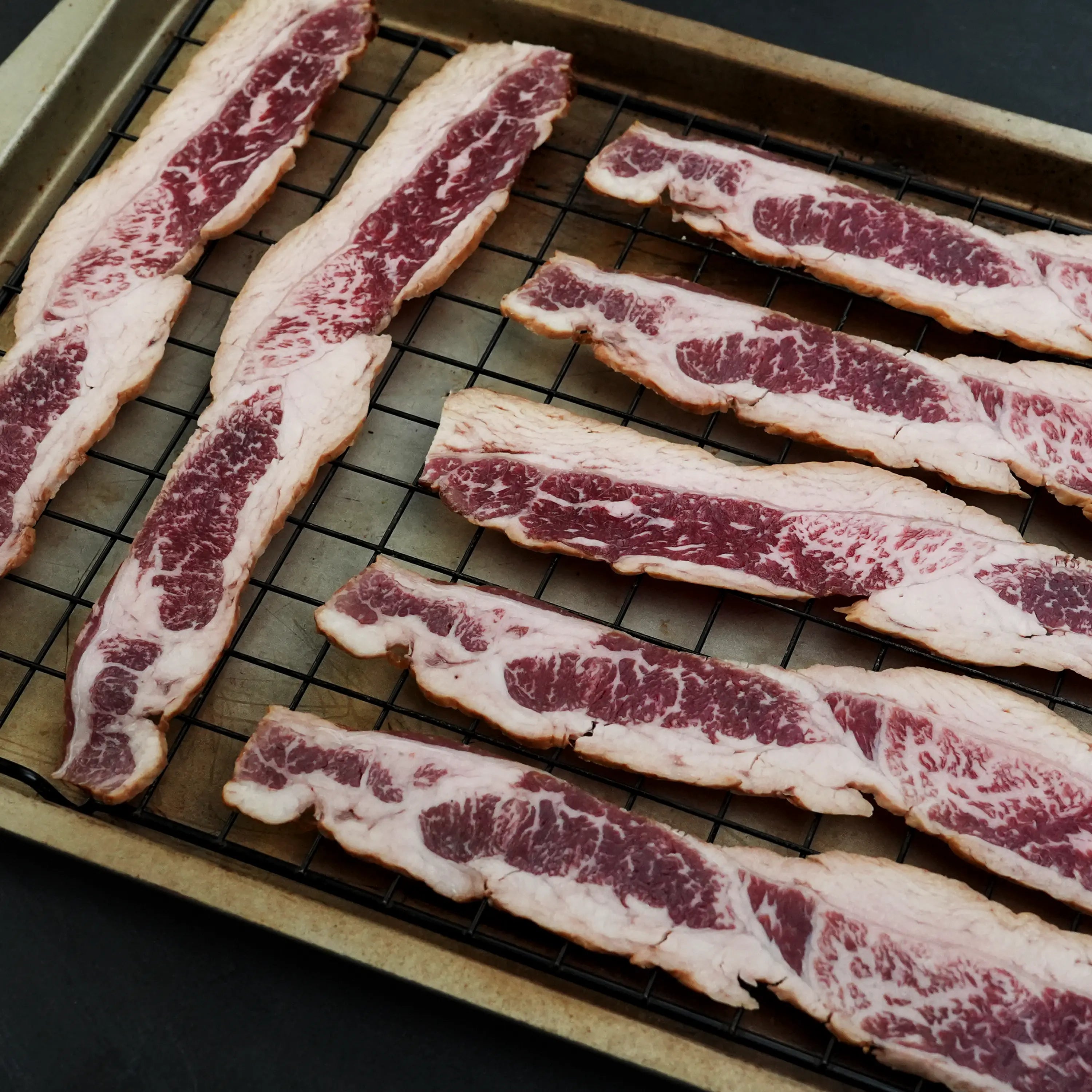 Westholme Australian Wagyu Halal Beef Bacon slices, richly marbled and laid on a wire rack, showcasing their cured and smoked, beefy excellence.