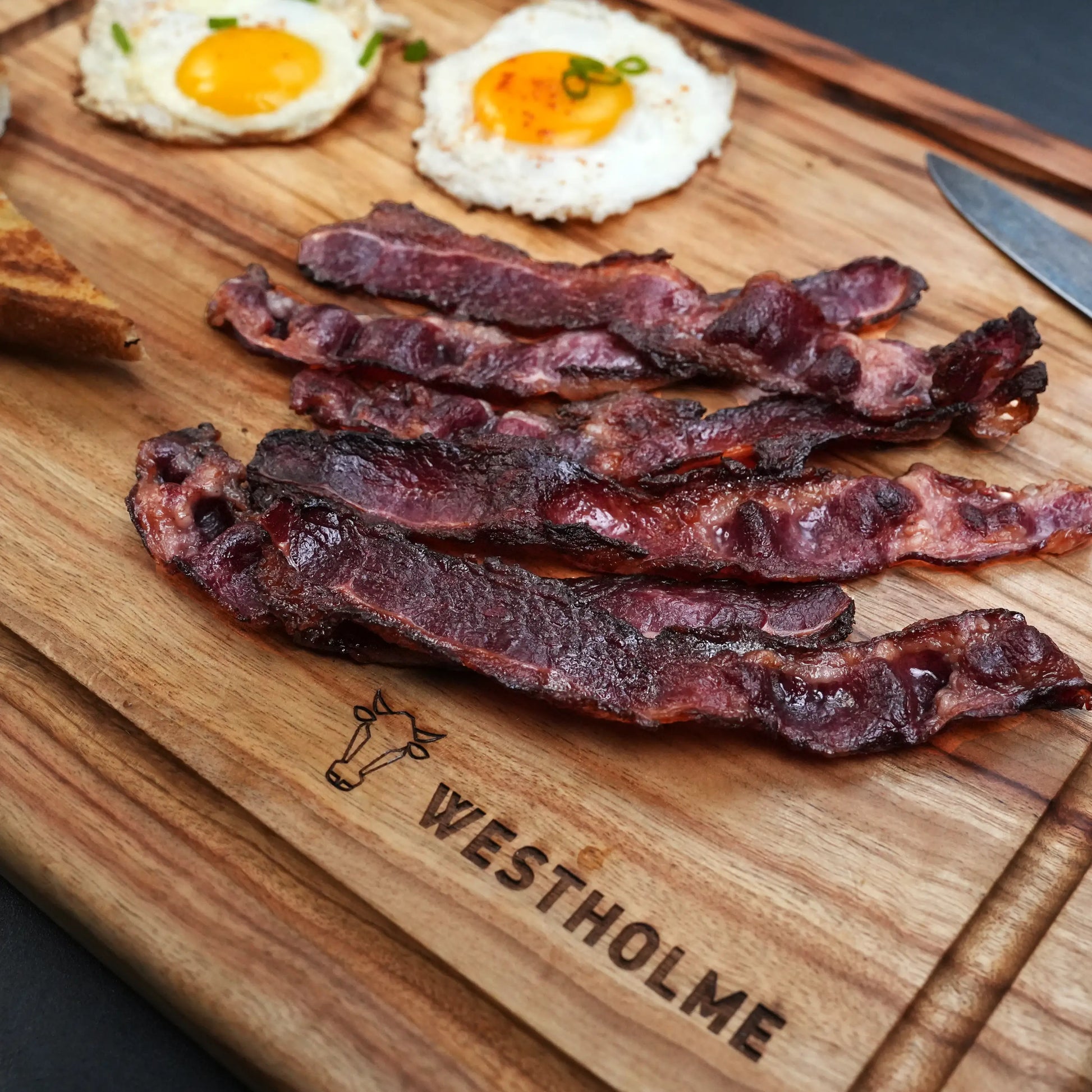 Westholme Australian Wagyu Halal Beef Bacon on a wooden board with two sunny-side-up eggs and toast, showcasing its rich, smoky flavor.
