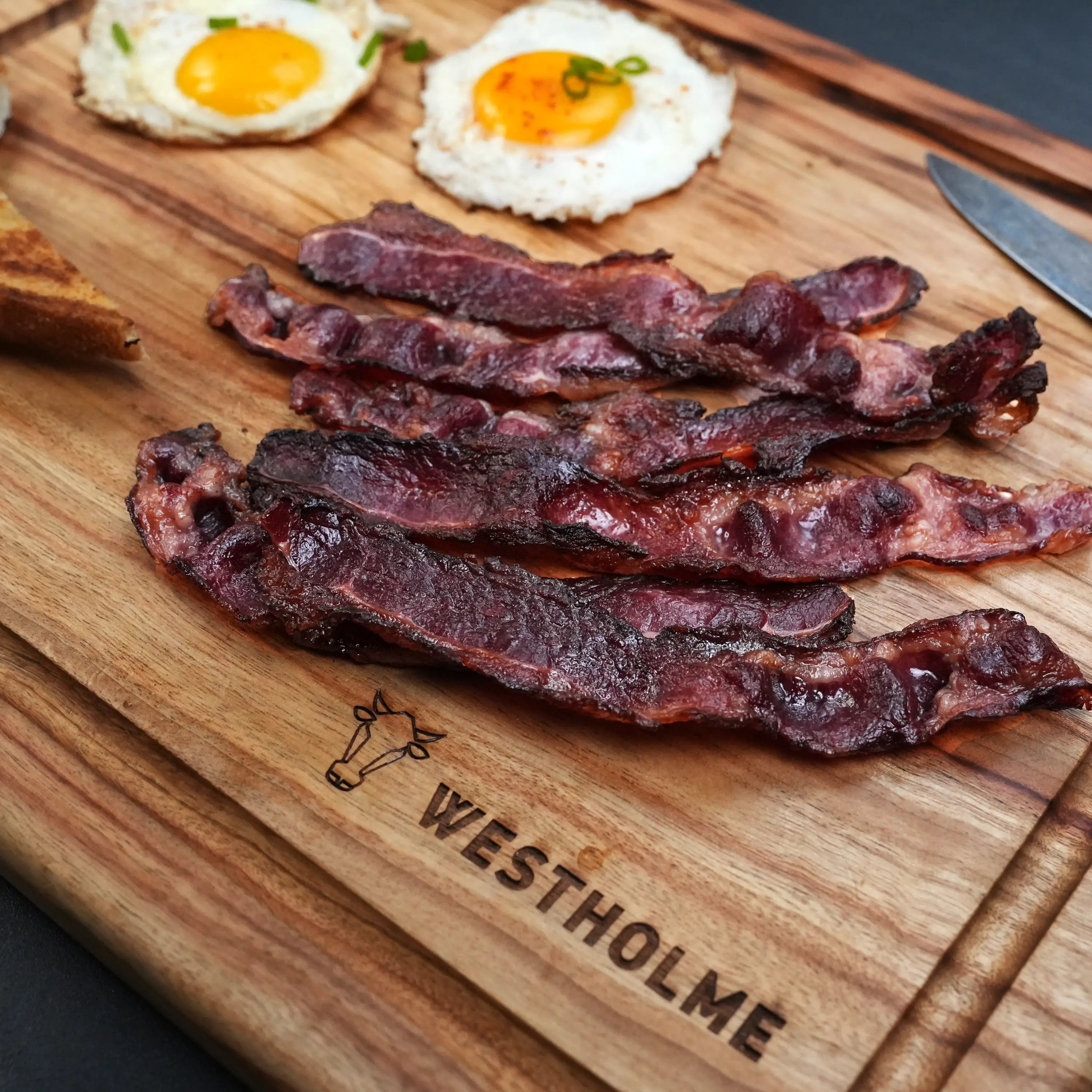 Westholme Australian Wagyu Halal Beef Bacon on a wooden board with two sunny-side-up eggs and toast, showcasing its rich, smoky flavor.