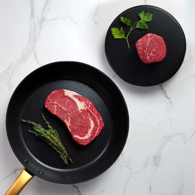 King & Queen Choice Angus Combo: Rib-eye in black pan with thyme, filet mignon on plate with parsley, showcasing premium, marbled Black Angus steaks.