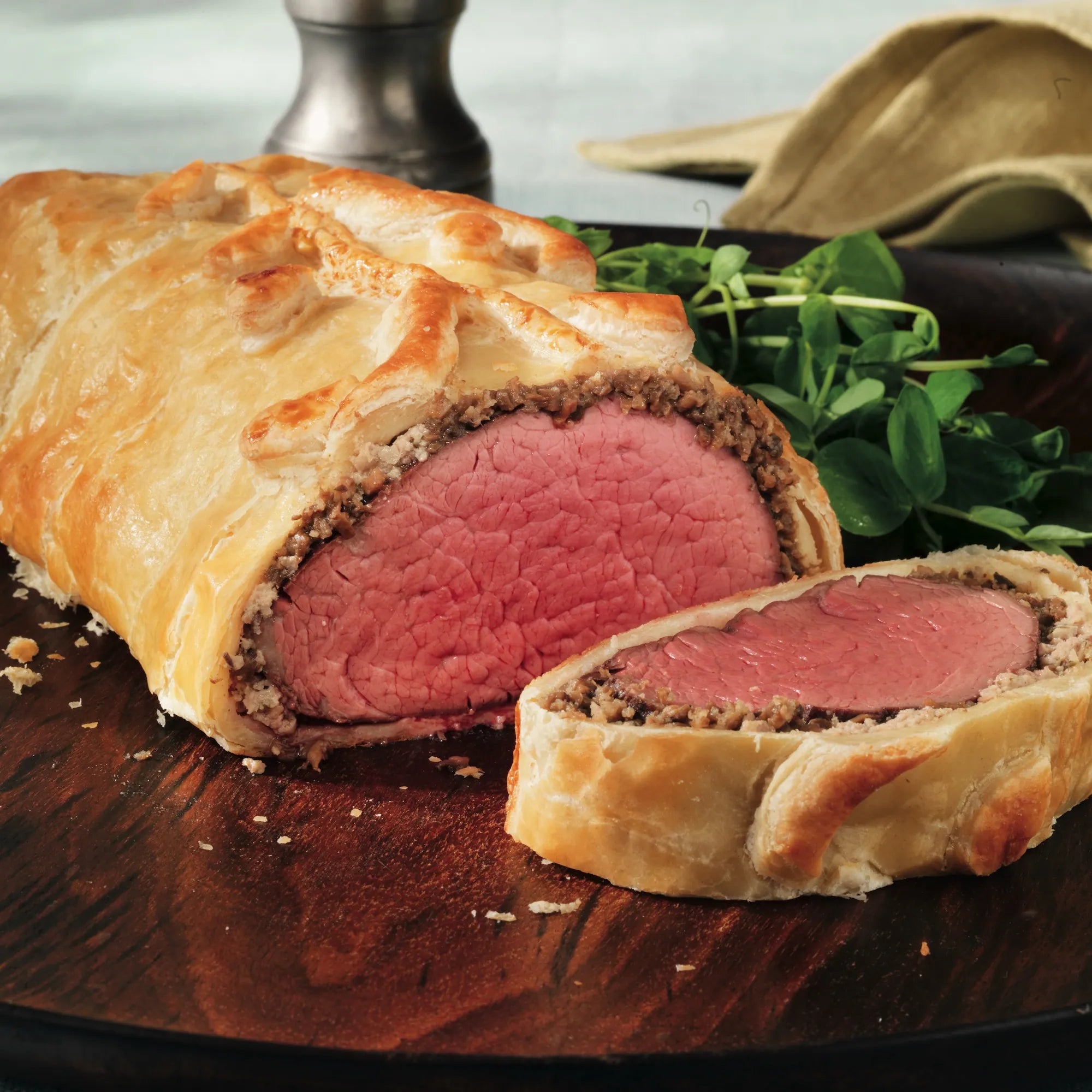 Roast Sized Beef Wellington with medium-rare beef tenderloin in mushroom duxelles, wrapped in golden puff pastry, sliced on a wooden board, garnished with greens.